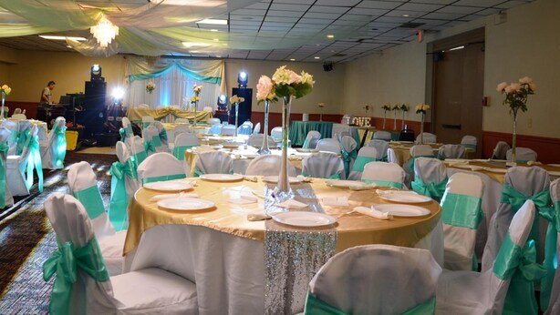 Un salón de banquetes con mesas redondas dispuestas para comer, sillas con cintas verdes y centros de mesa florales.