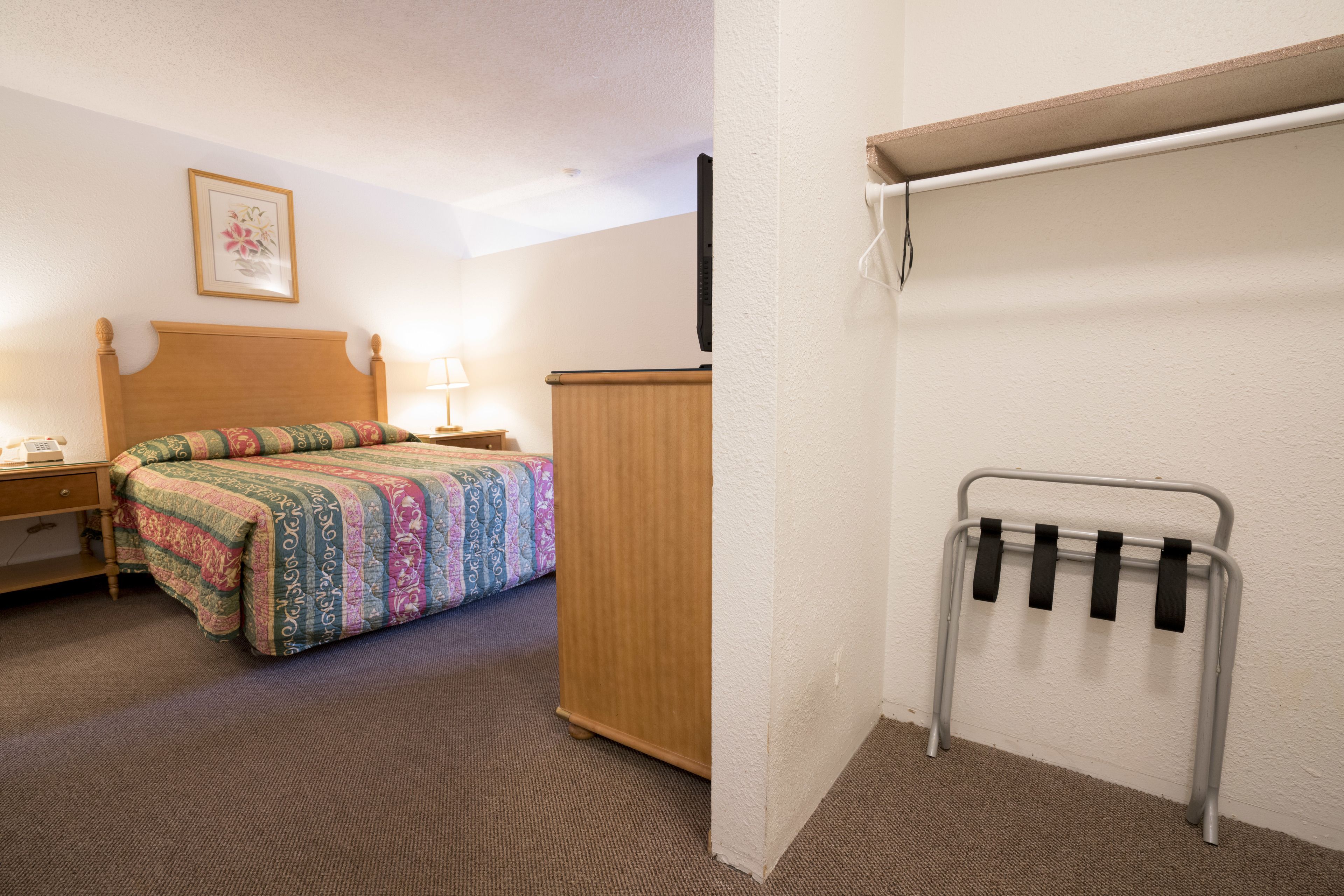 A hotel room with a bed, a wooden wardrobe, a bedside table with a lamp, and a wall-mounted towel rack.