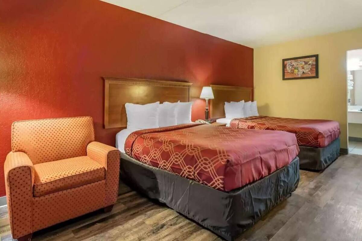 A hotel room with two beds, a sofa, a wooden headboard, a lamp, and a framed picture on the wall.