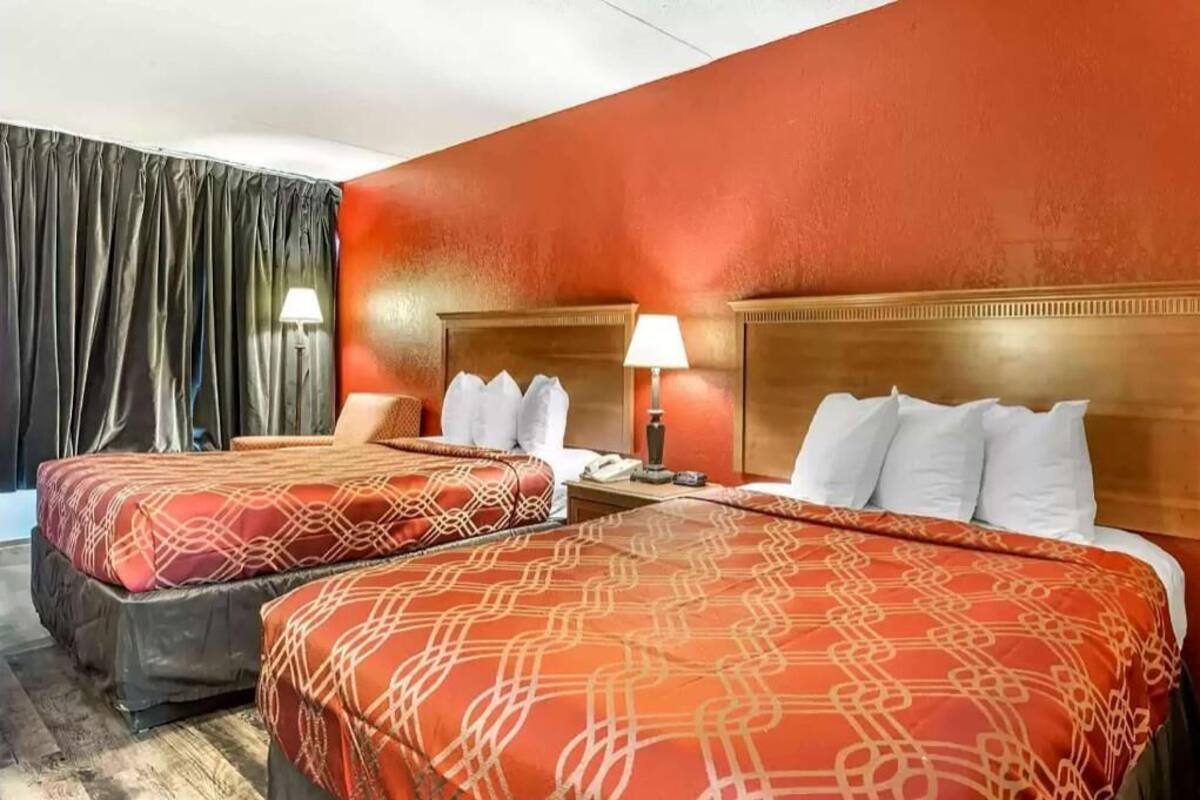 A hotel room with two beds, a wooden headboard, a nightstand with a lamp, and a curtain.