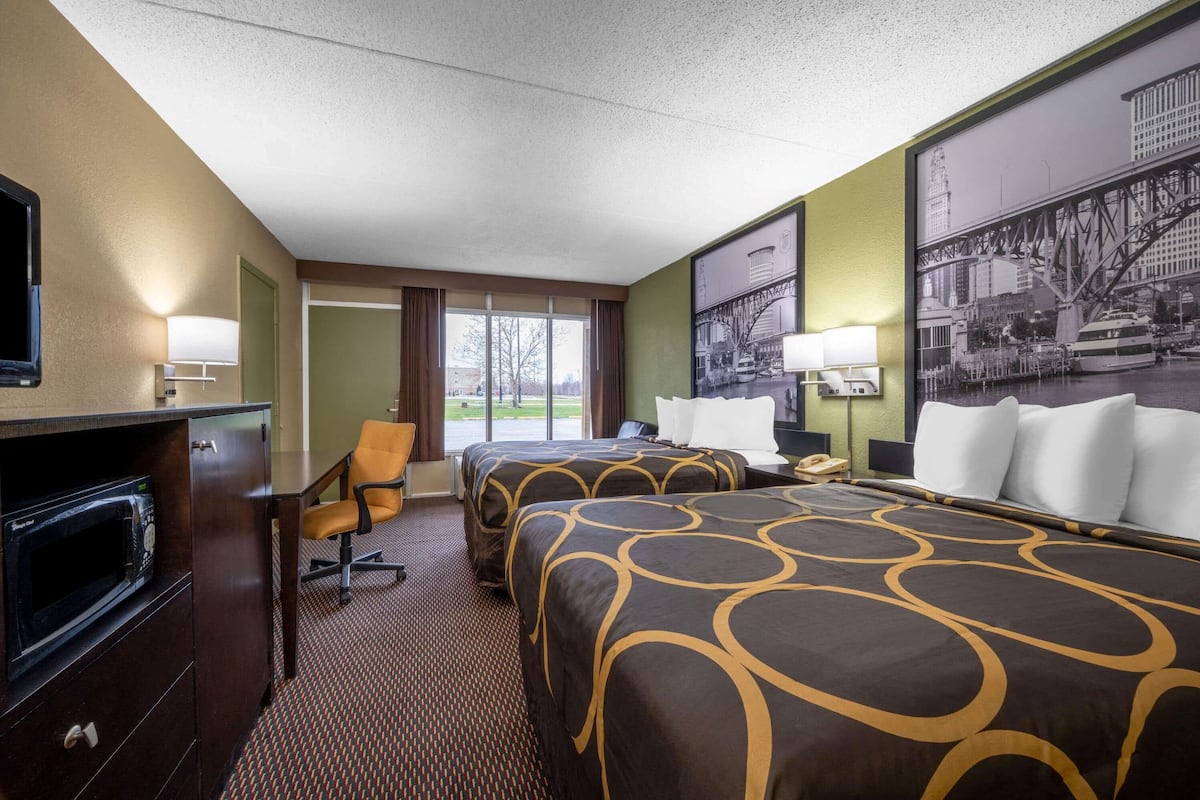 A hotel room with two beds, a desk, a chair, a microwave, and a large window.