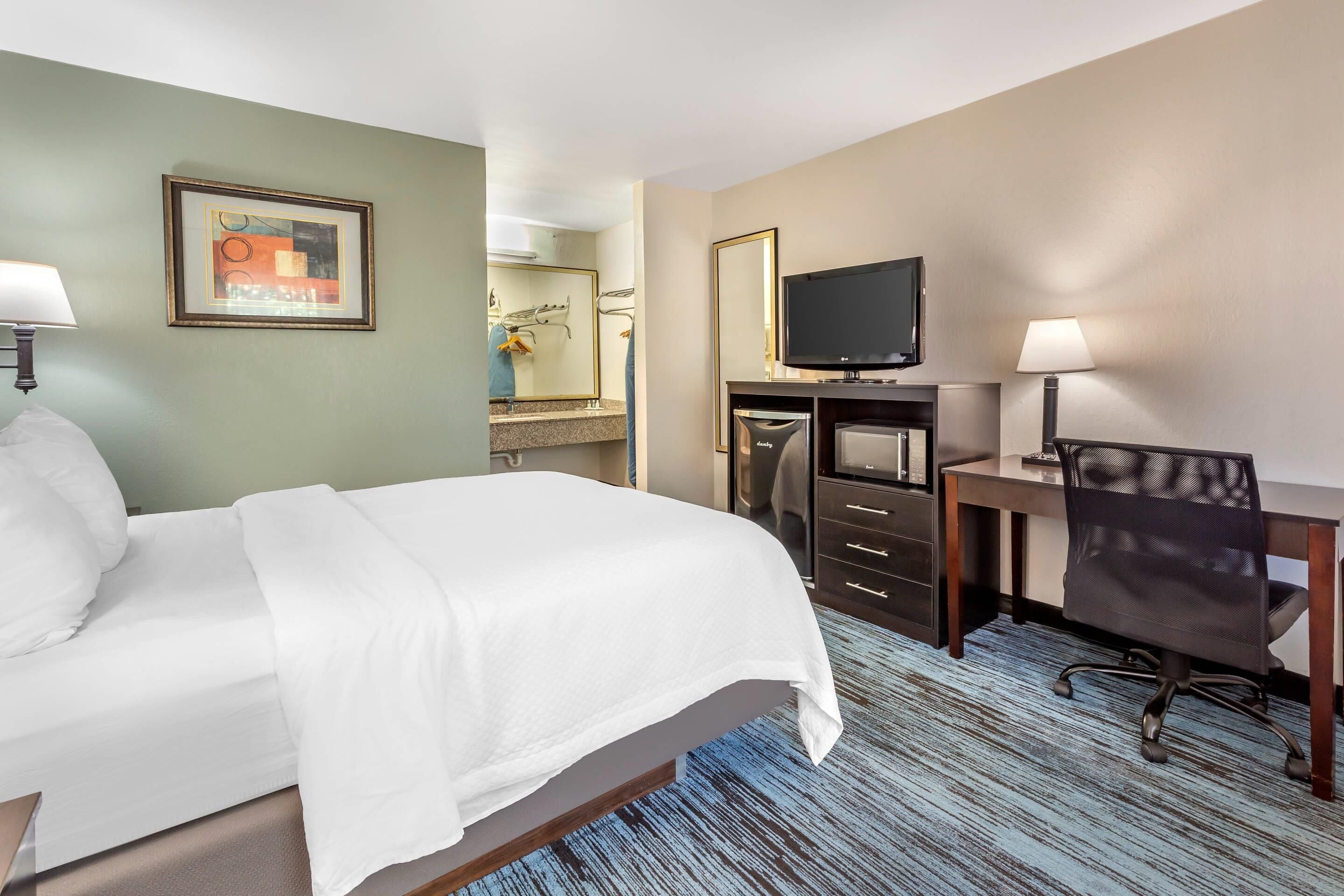 A hotel room with a bed, a desk, a chair, a television, and a bathroom visible in the background.