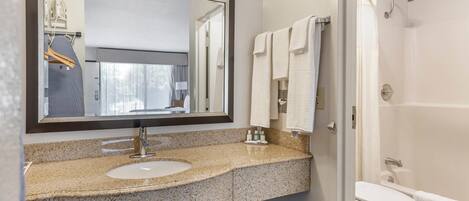 Baño con lavamanos de encimera de granito, un espejo grande, toallero y zona de ducha.