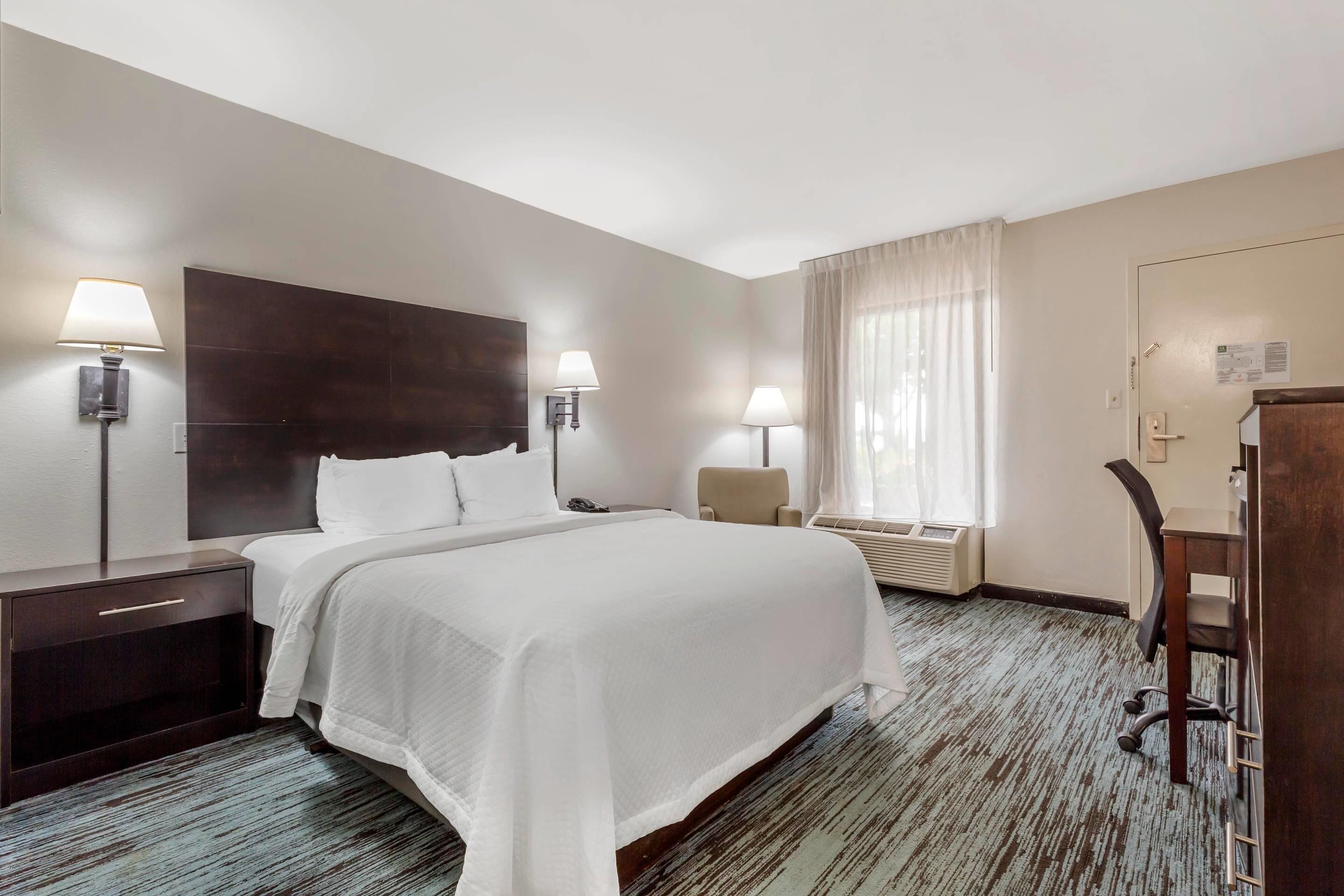 A hotel room with a large bed, two bedside tables with lamps, a desk with a chair, and a window with curtains.
