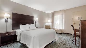 A hotel room with a large bed, two bedside tables with lamps, a desk with a chair, and a window with curtains.