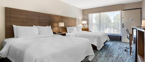 A hotel room with two beds, a large wooden headboard, a desk with a chair, and a large window with curtains.