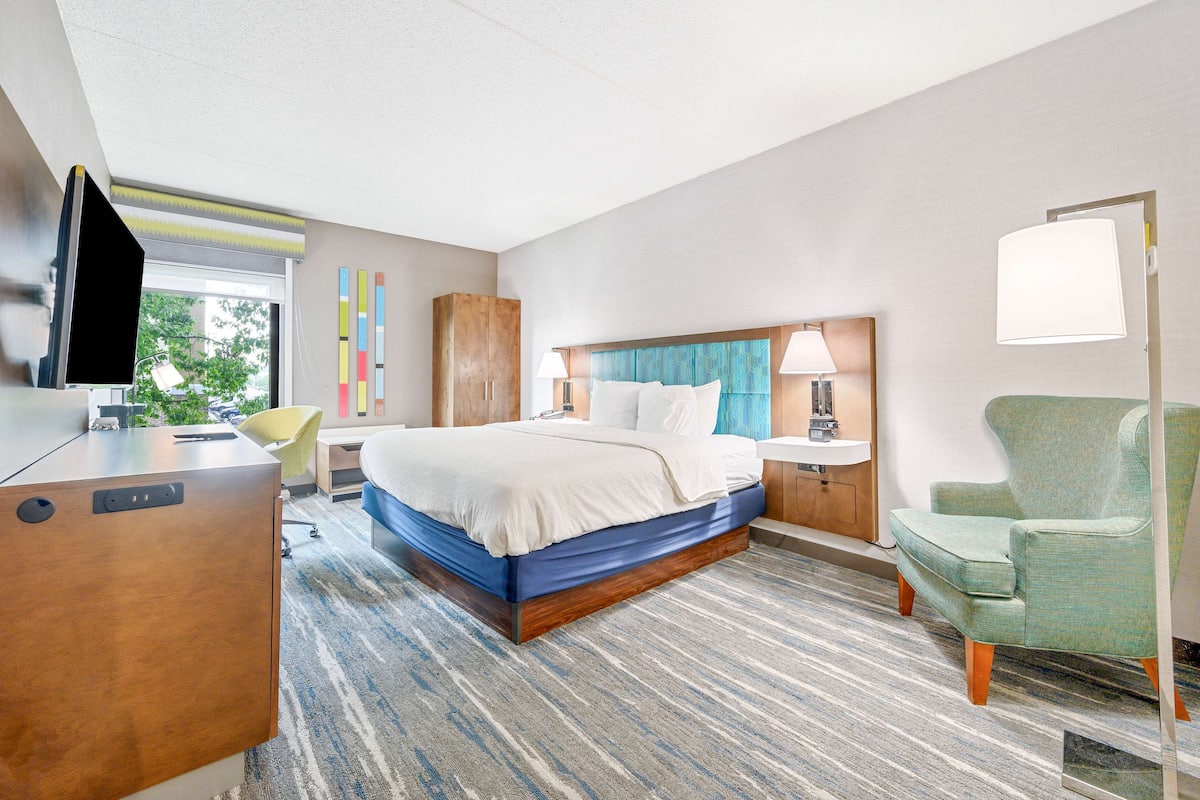 A hotel room with a bed, a desk, a chair, a lamp, and a window.