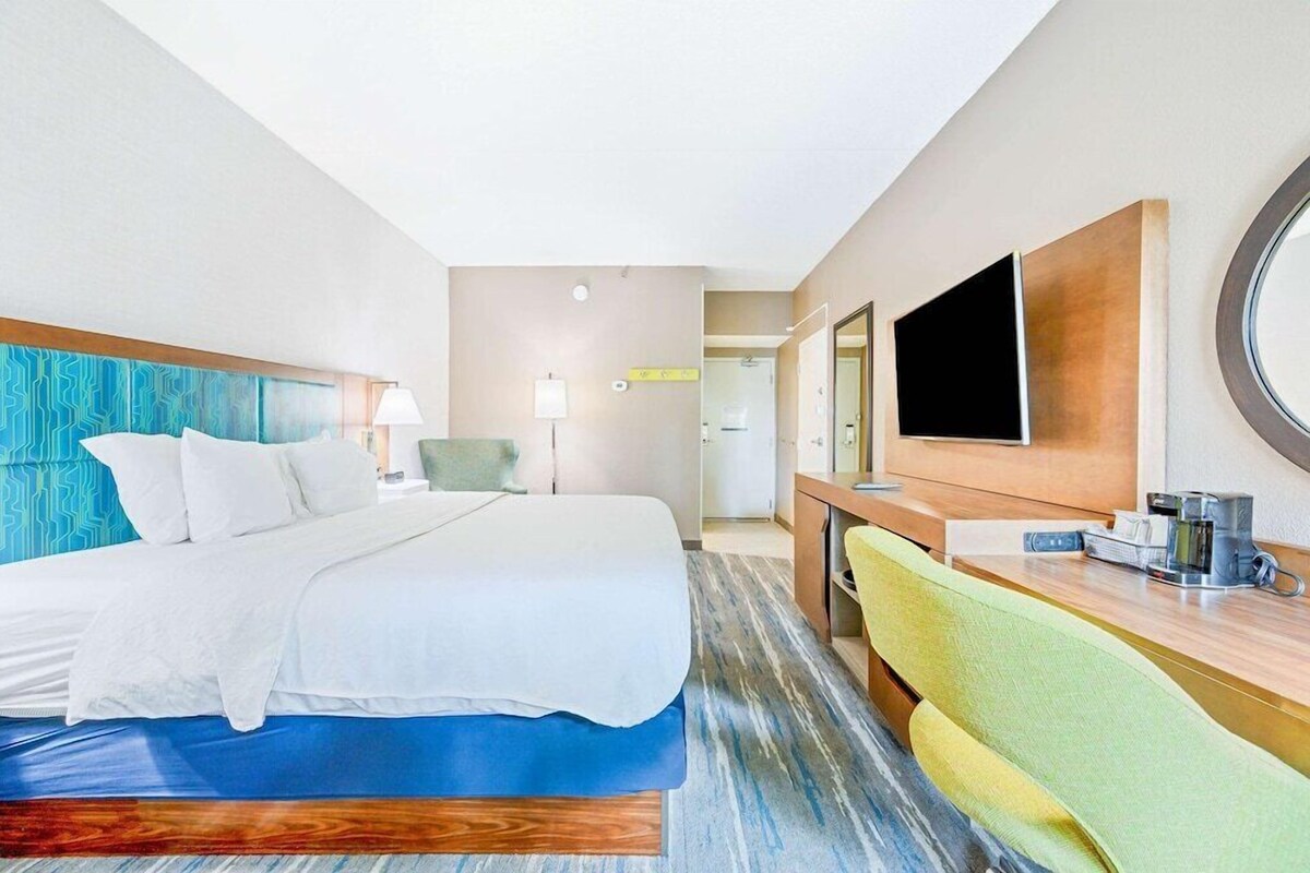 Une chambre d’hôtel avec un grand lit, un bureau avec une chaise, une télévision et une salle de bain visible au fond.