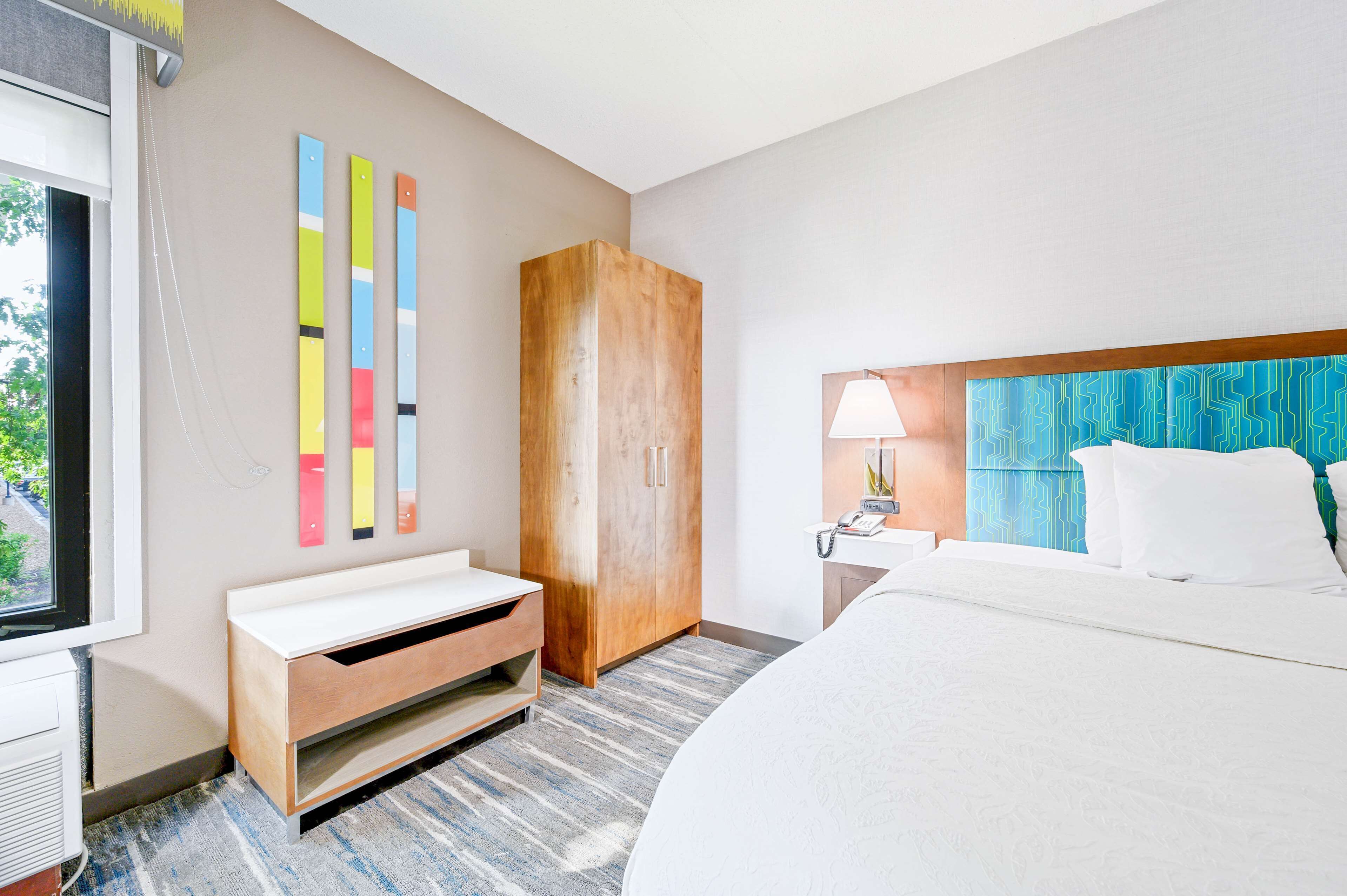 A hotel room with a bed, a wooden wardrobe, a bench, a nightstand, a lamp, and a wall with colorful vertical stripes.