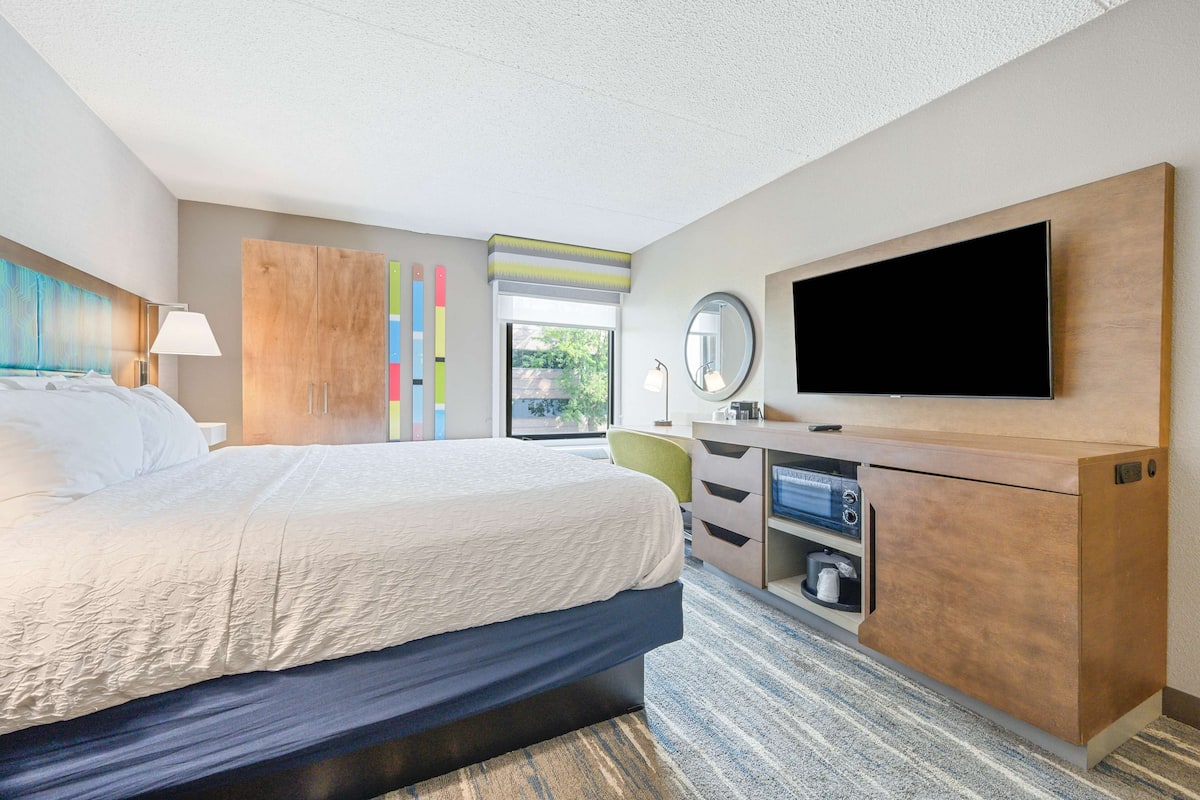 A modern hotel room with a large bed, a desk with a flat-screen TV, a mirror, and a window with a view of greenery.