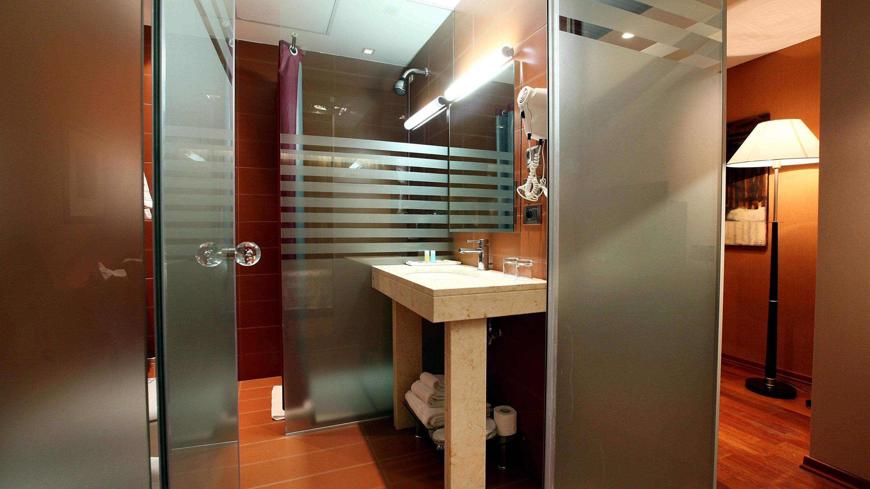 A modern bathroom with a glass shower enclosure, a sink with a mirror, and a standing lamp.