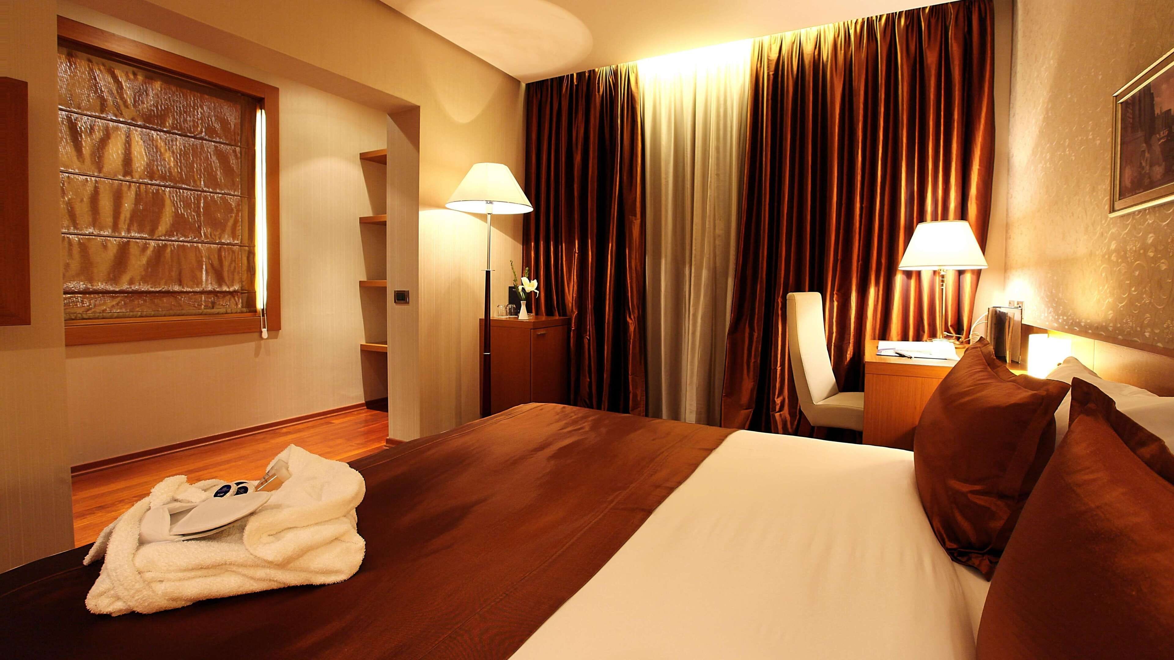 A hotel room with a large bed, a wooden floor, a desk with a lamp, a chair, and a window with curtains.