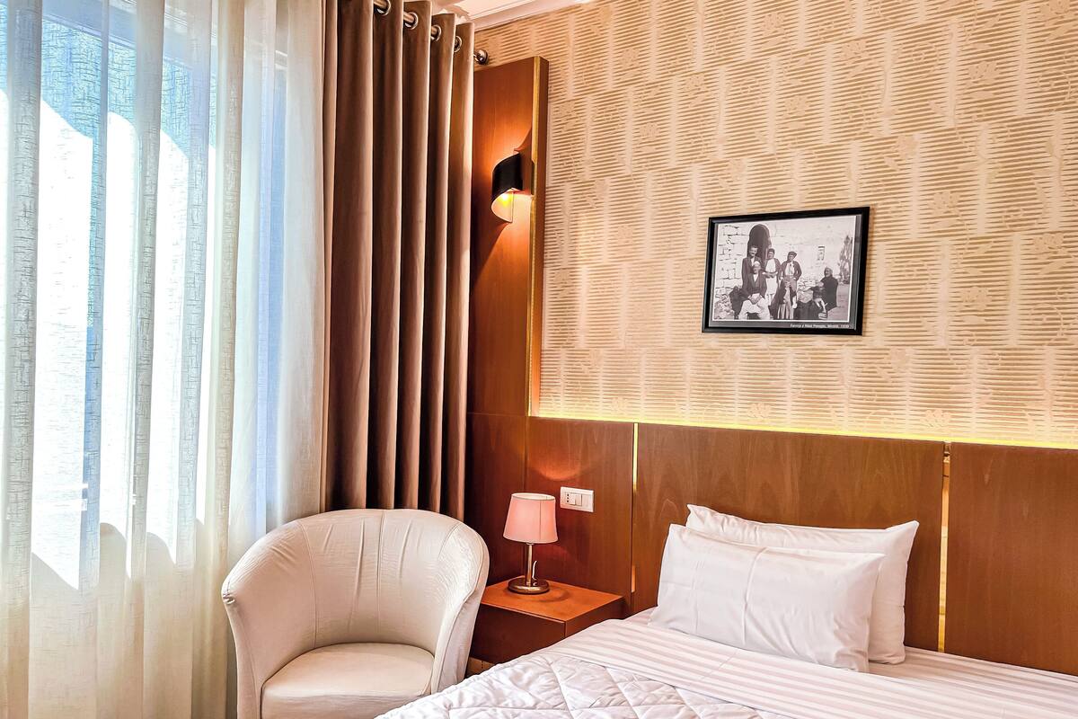 Hotellihuone, jossa on sänky, tuoli, yöpöytä, lamppu ja seinällä oleva kehystetty kuva.