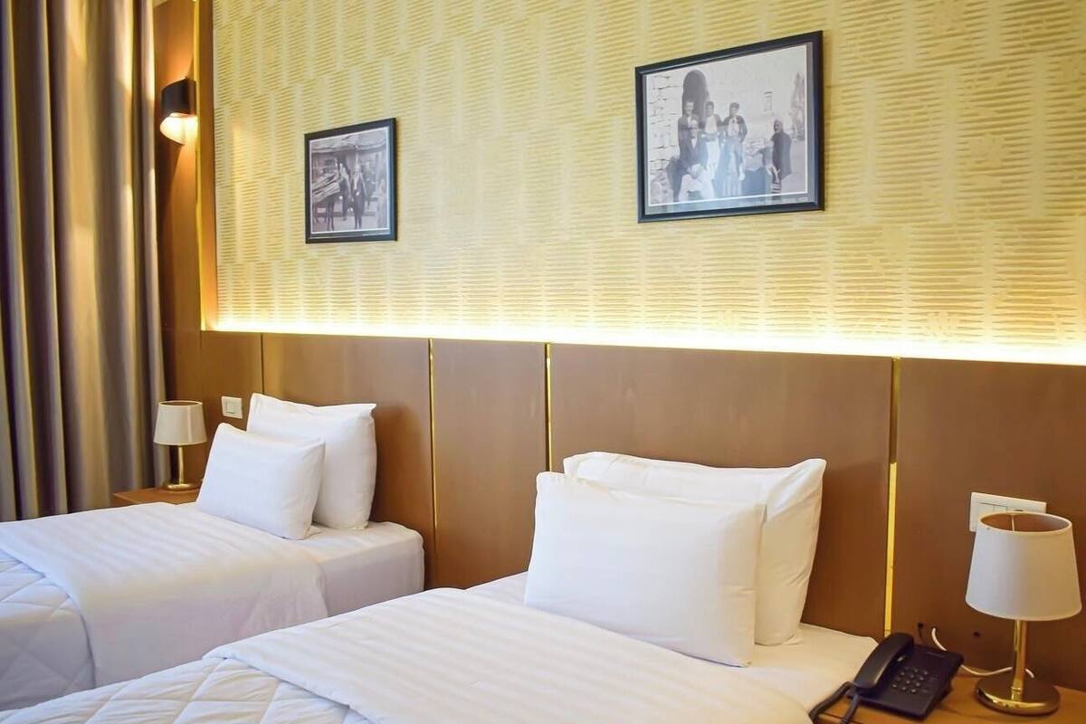 Hotellihuone, jossa on kaksi sänkyä, yöpöytä lampun kanssa, puhelin ja seinällä kehystettyjä valokuvia.
