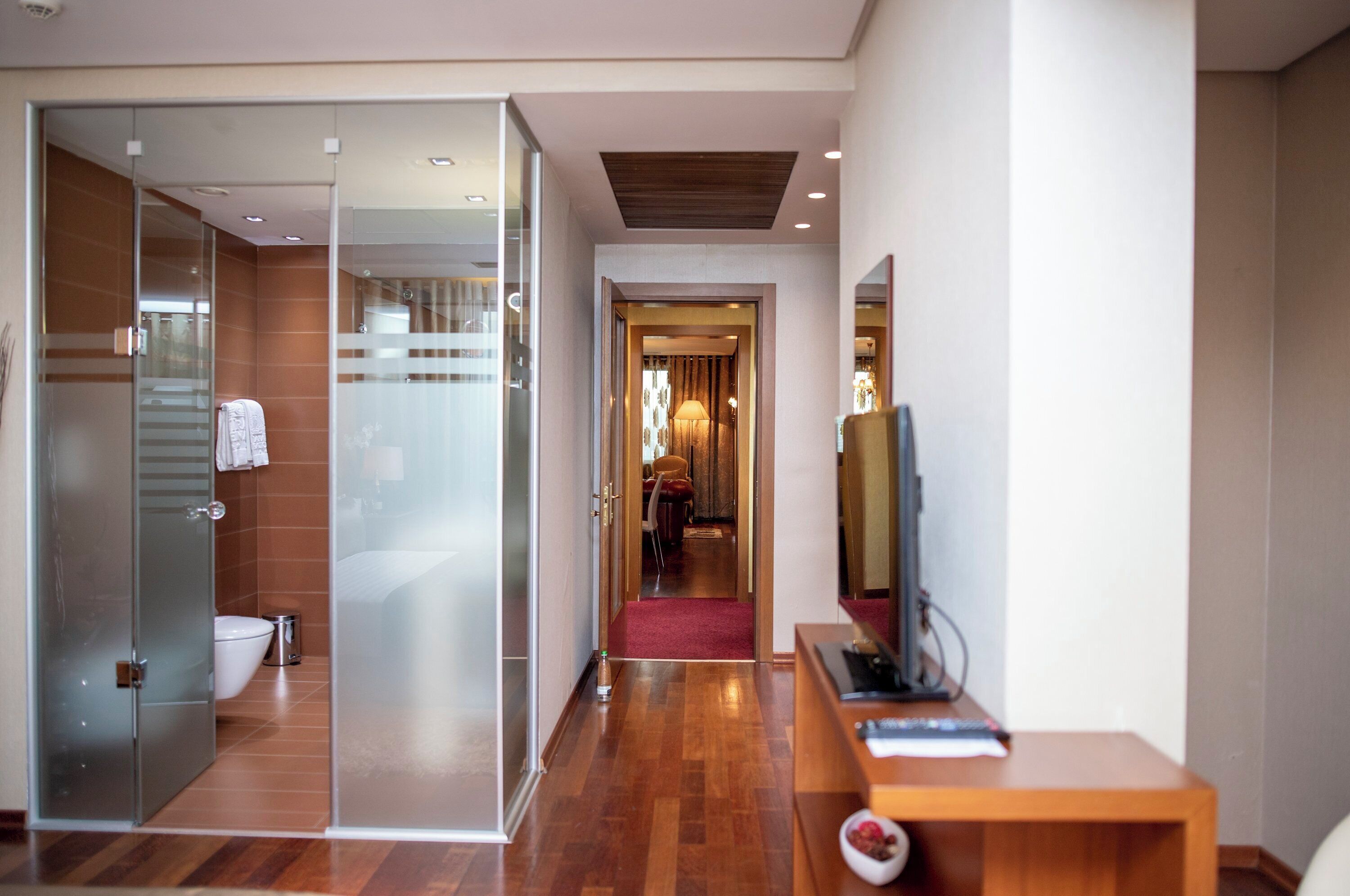 A modern hotel room with a glass shower enclosure, a wooden floor, a desk with a computer, and a television.
