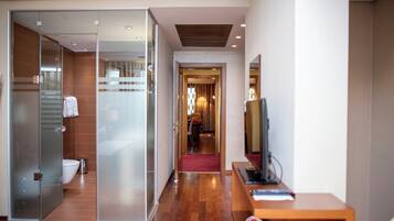 A modern hotel room with a glass shower enclosure, a wooden floor, a desk with a computer, and a television.