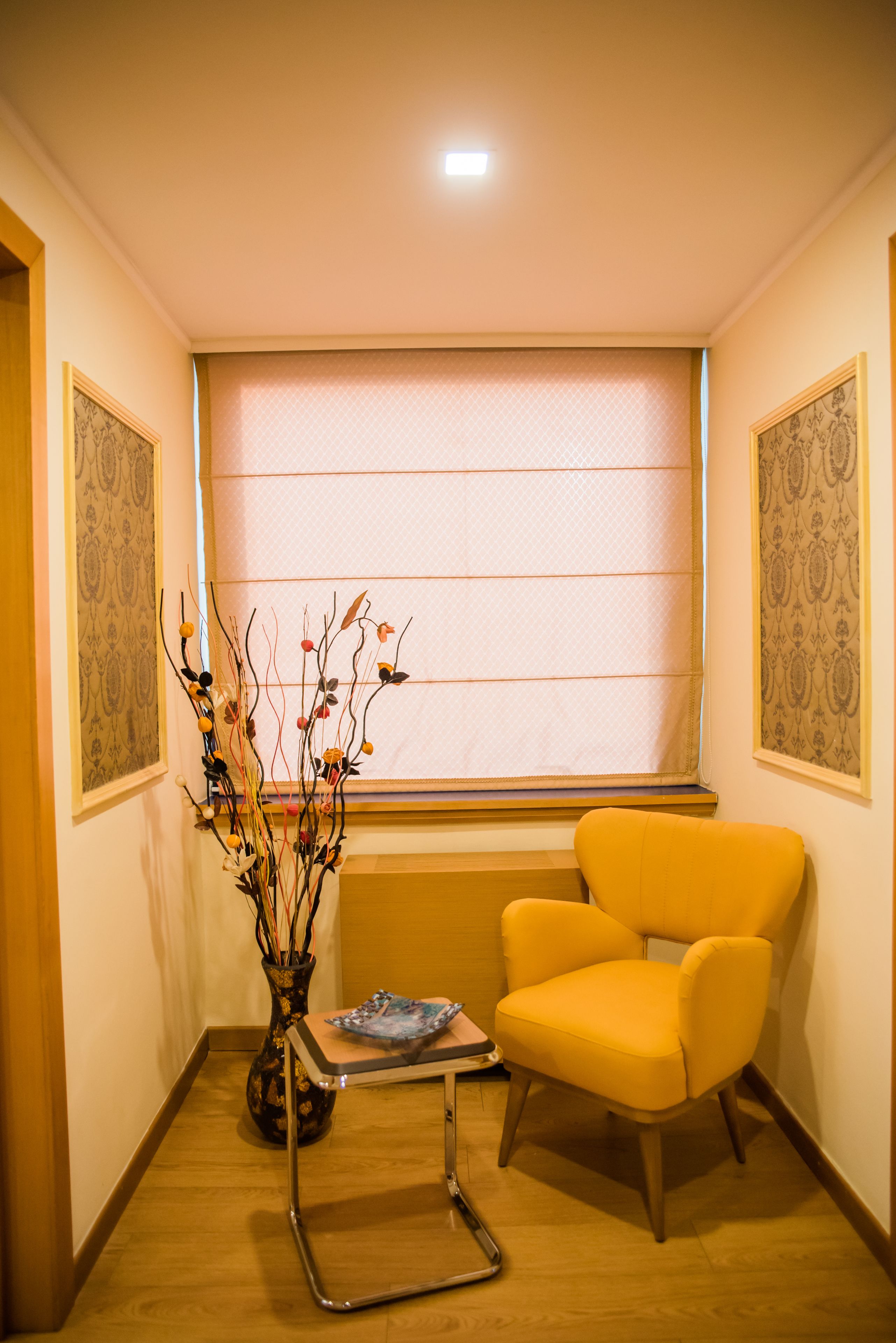 Una habitación de esquina con un sillón amarillo, una mesita, un florero con ramas y un papel tapiz estampado.