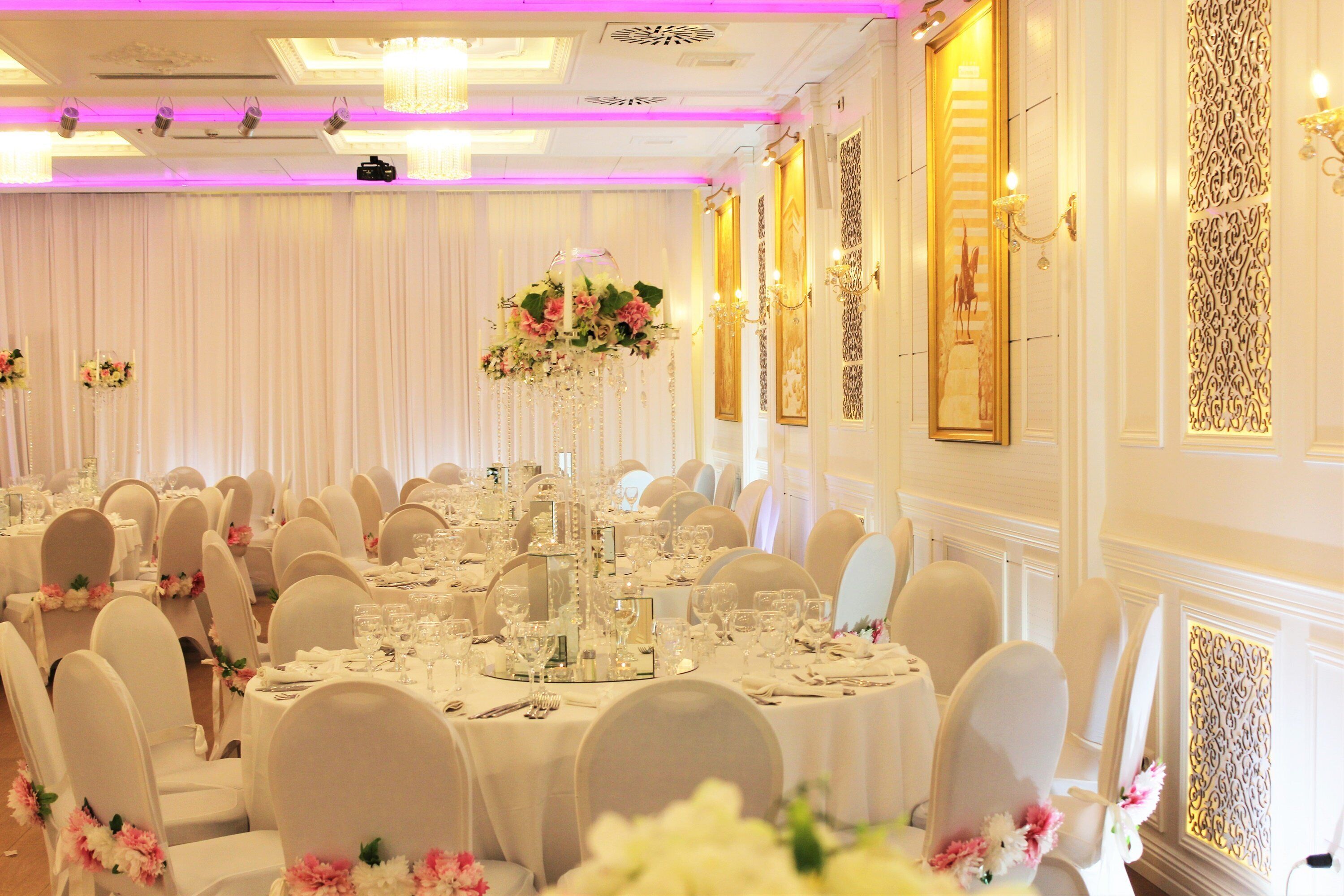 Un salón de banquetes con mesas redondas dispuestas para cenar, sillas blancas y centros de mesa florales.