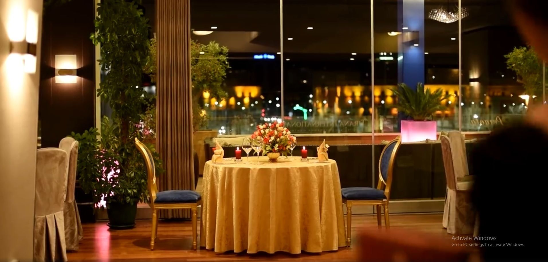 A romantic dinner setting with a table set for two, surrounded by chairs, a potted plant, and a view of the cityscape at night.