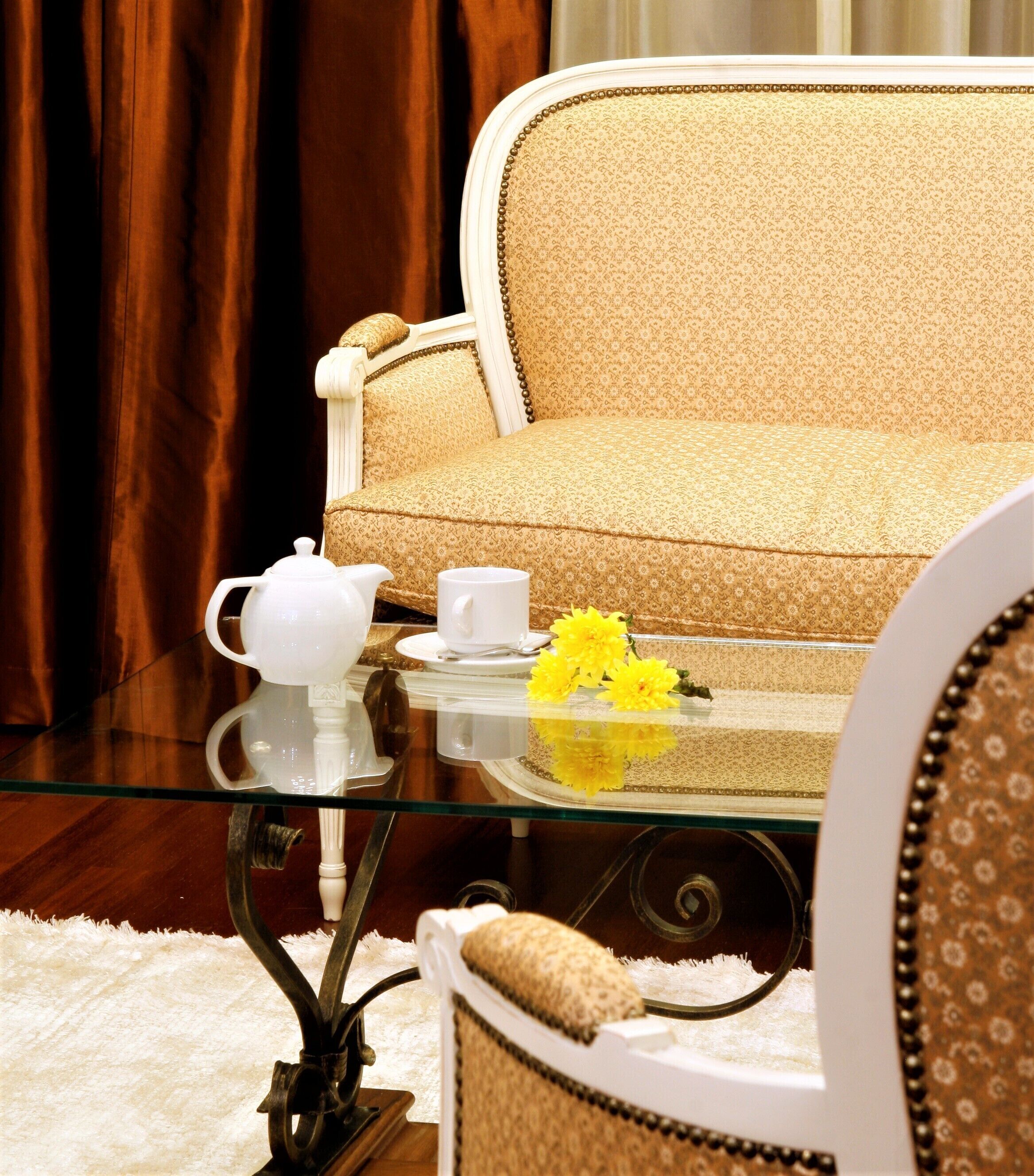 A sofa with a patterned fabric, a glass table with a teapot and cups, and a vase with yellow flowers.