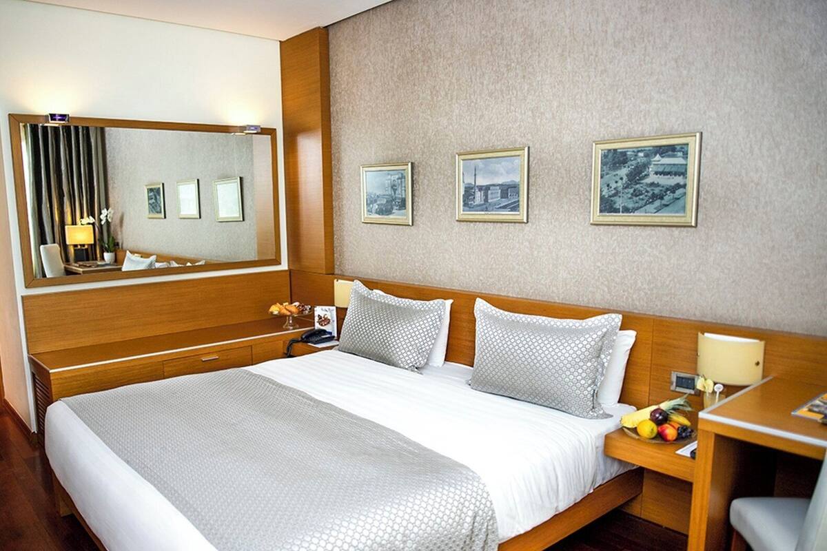 Hotellihuone, jossa on suuri sänky, puinen pääty, yöpöytä lampun kanssa ja kolme kehystettyä kuvaa seinällä.