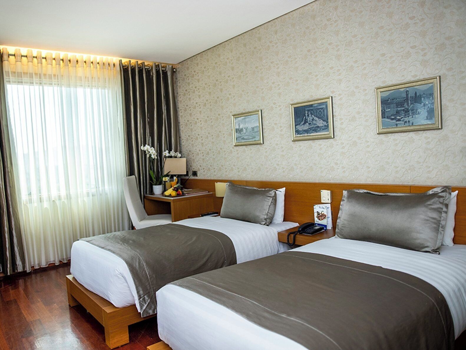 Habitación de hotel con dos camas, una mesita de noche, una lámpara, una pequeña mesa con una planta y tres cuadros en la pared.