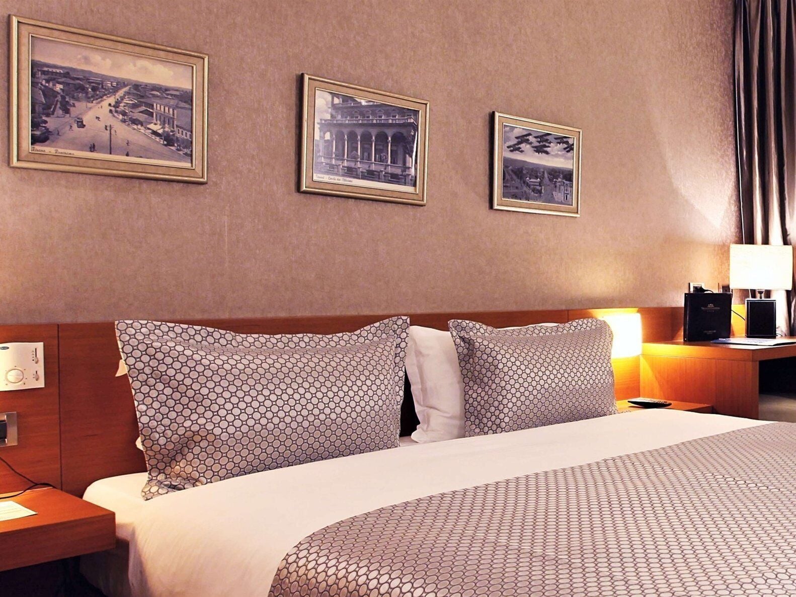 A hotel room with a large bed, two framed pictures on the wall, a nightstand with a lamp, and a wooden headboard.