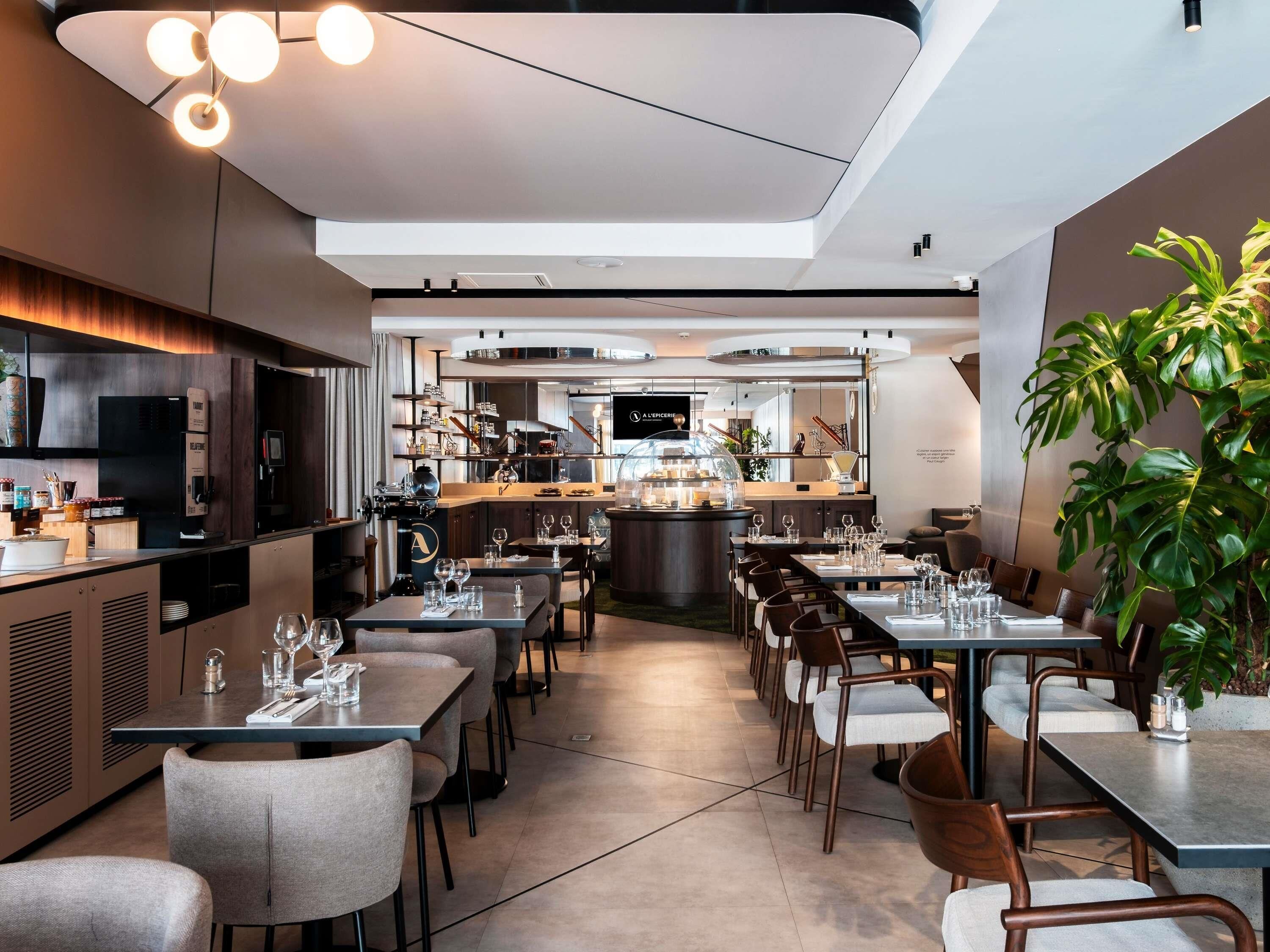 A modern restaurant interior with a long dining area, kitchen in view, and a large plant on the right.