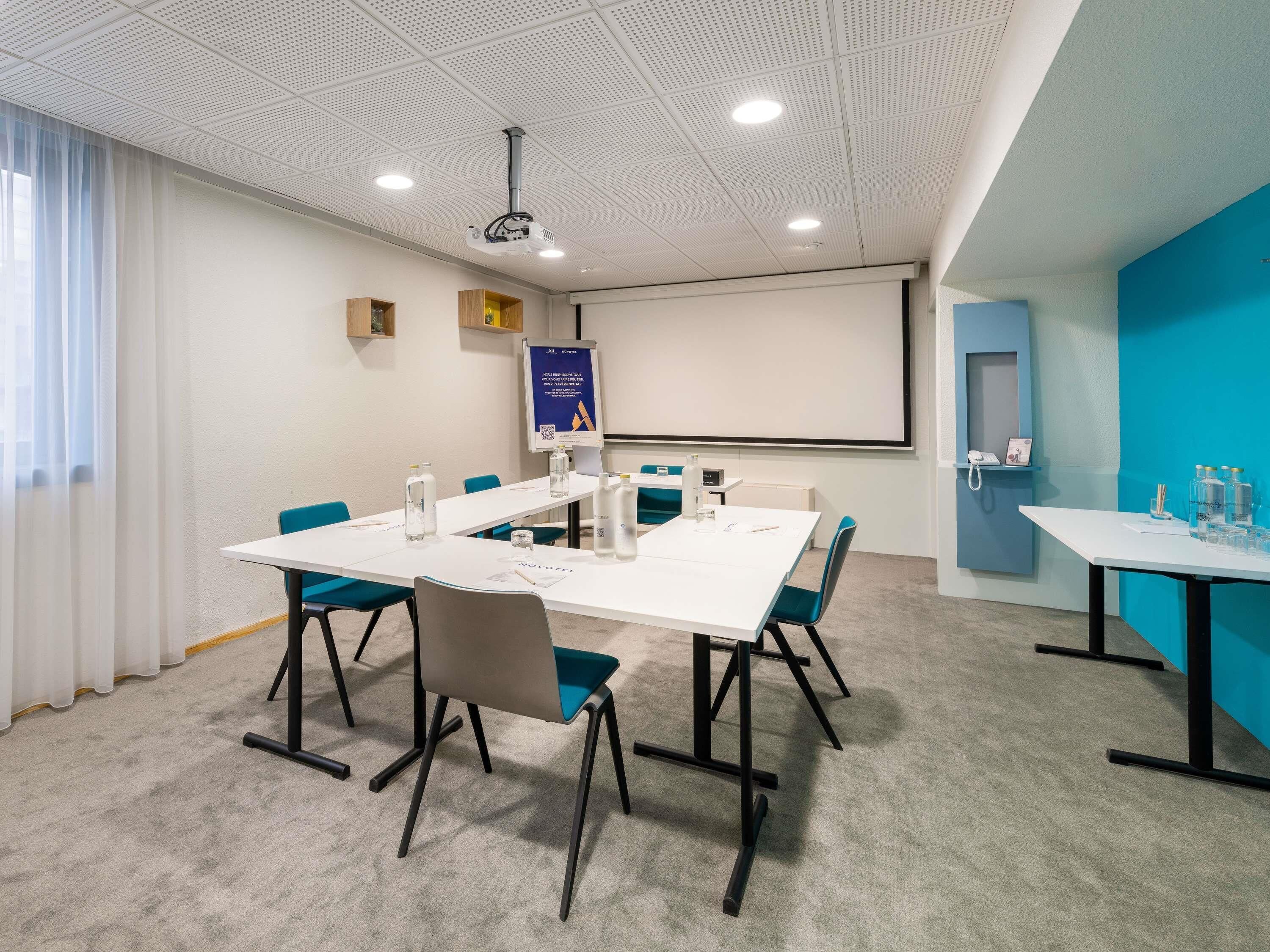 A conference room with a white table, teal chairs, a projector, and a screen.