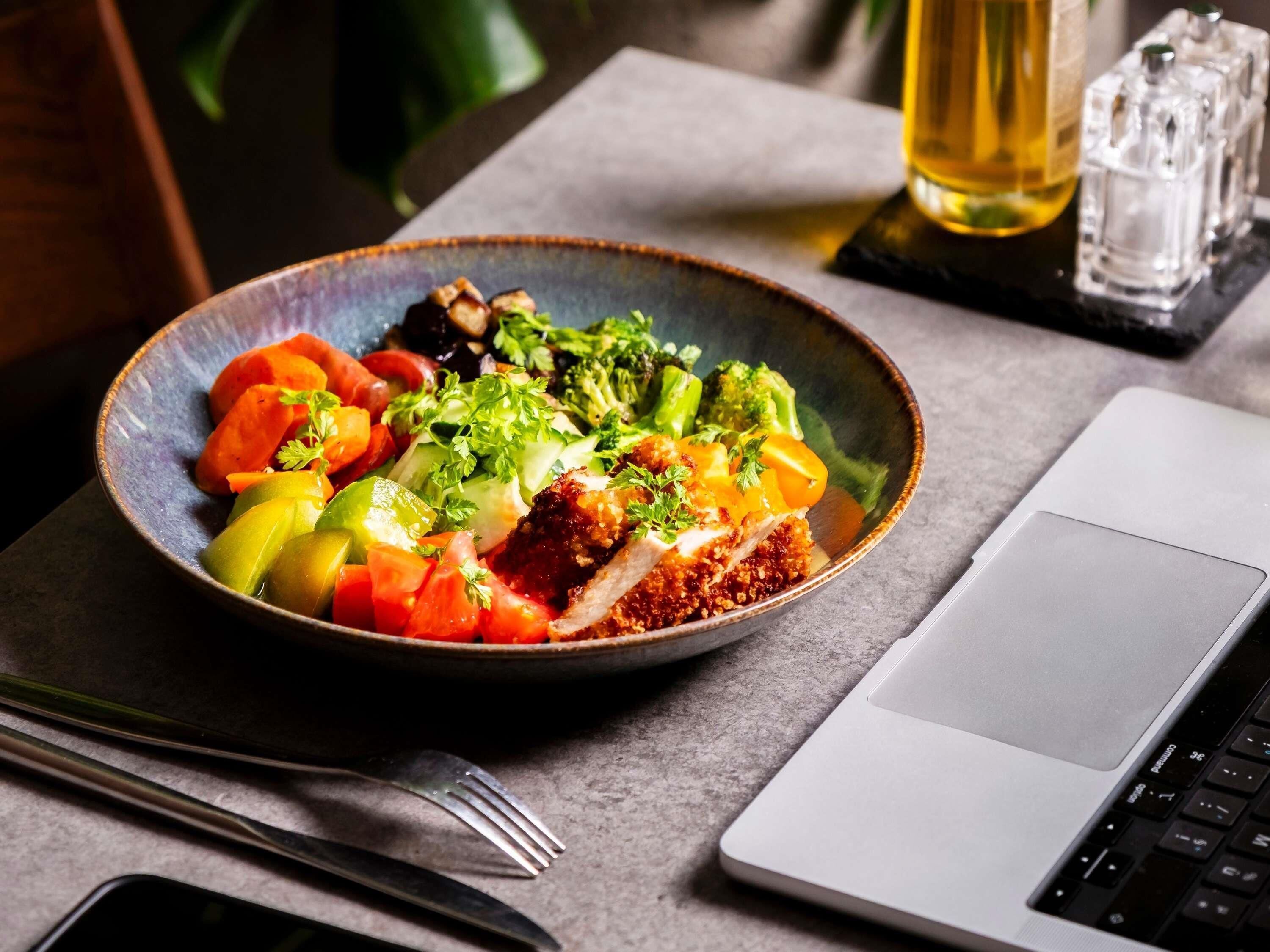 A bowl of food with a laptop and utensils on a table.