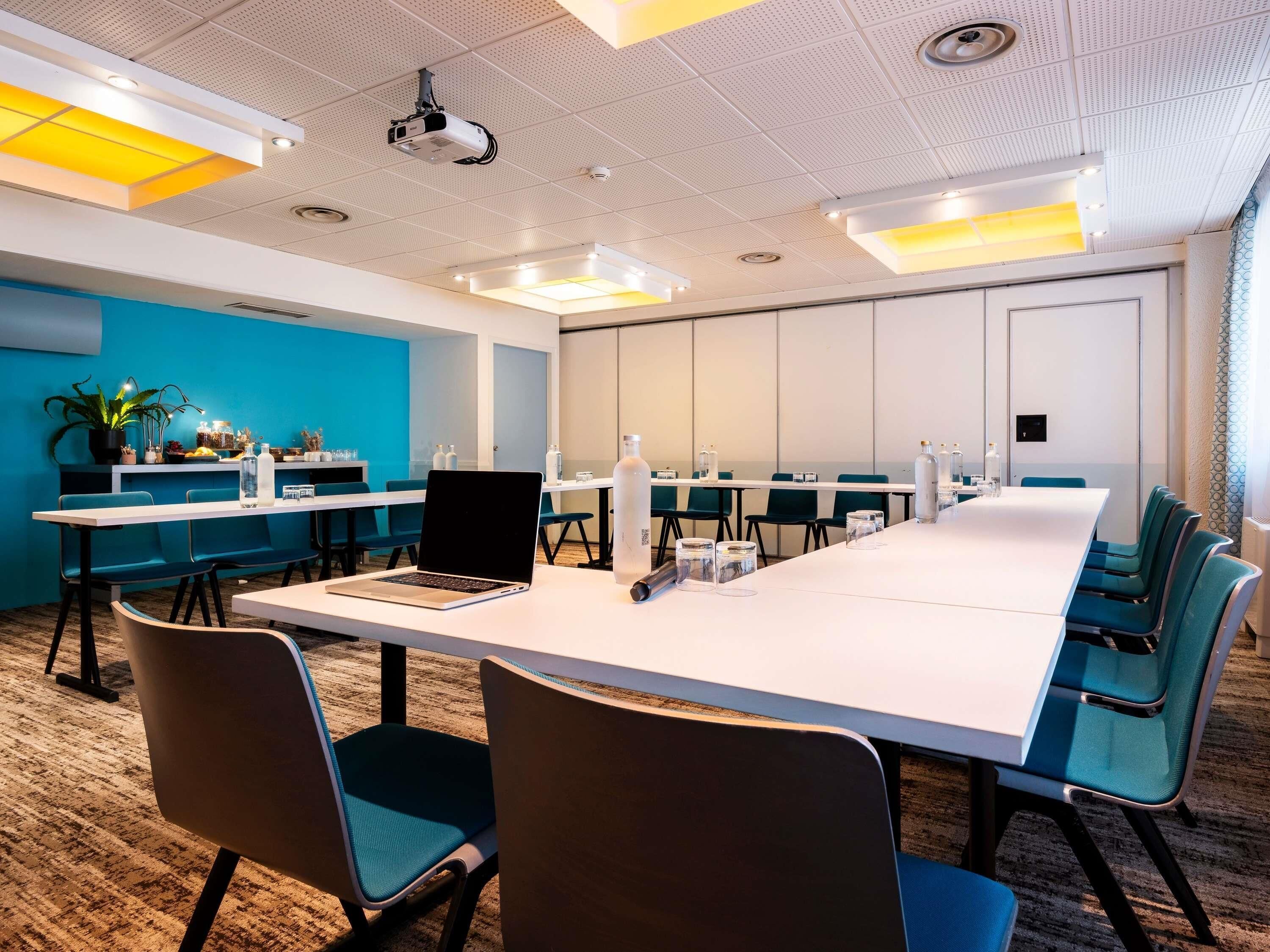A conference room with a long table, chairs, and a projector. There are plants and water bottles on the tables.