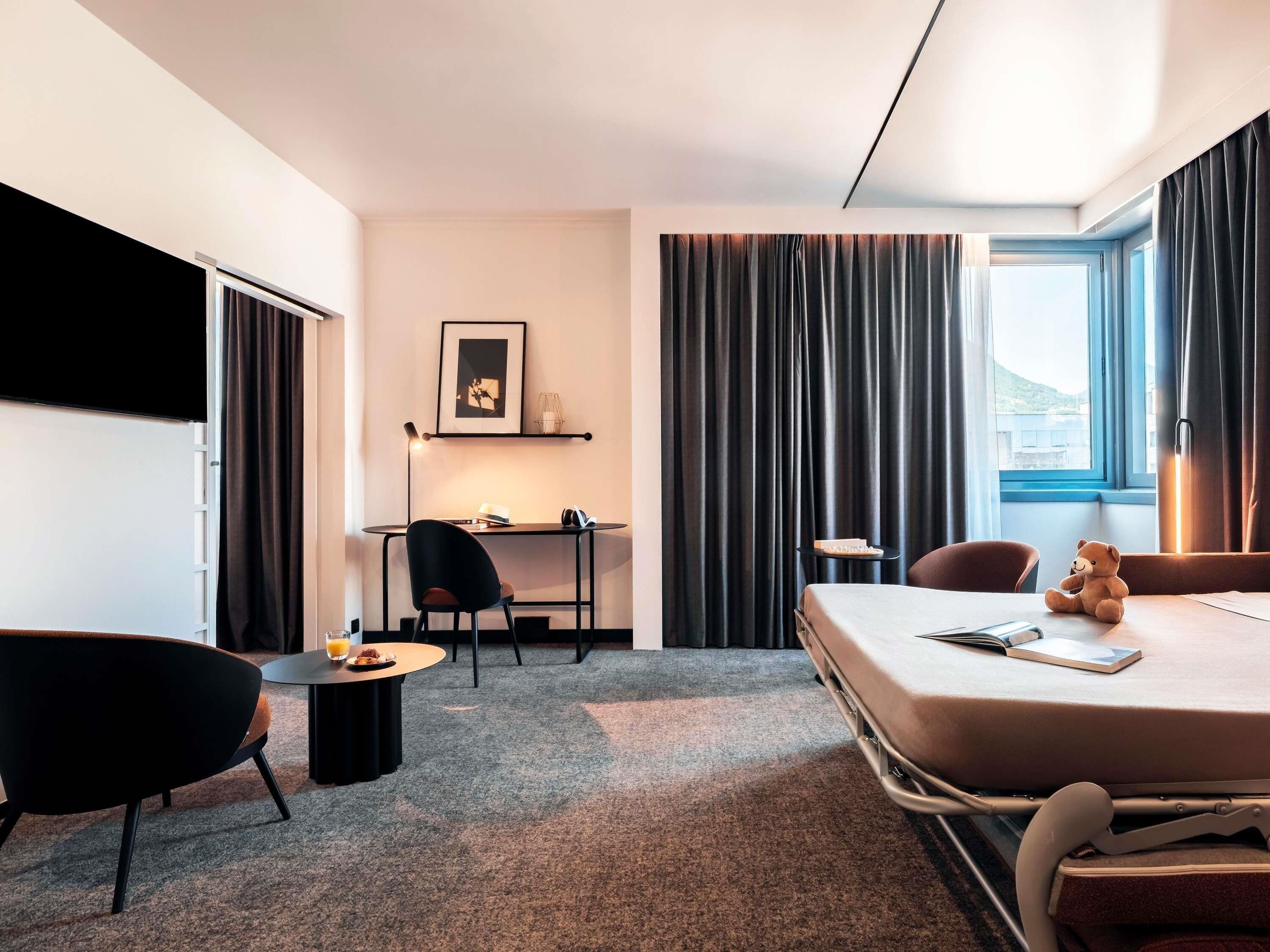 A hotel room with a bed, a desk, a chair, a small table, and a window with curtains.