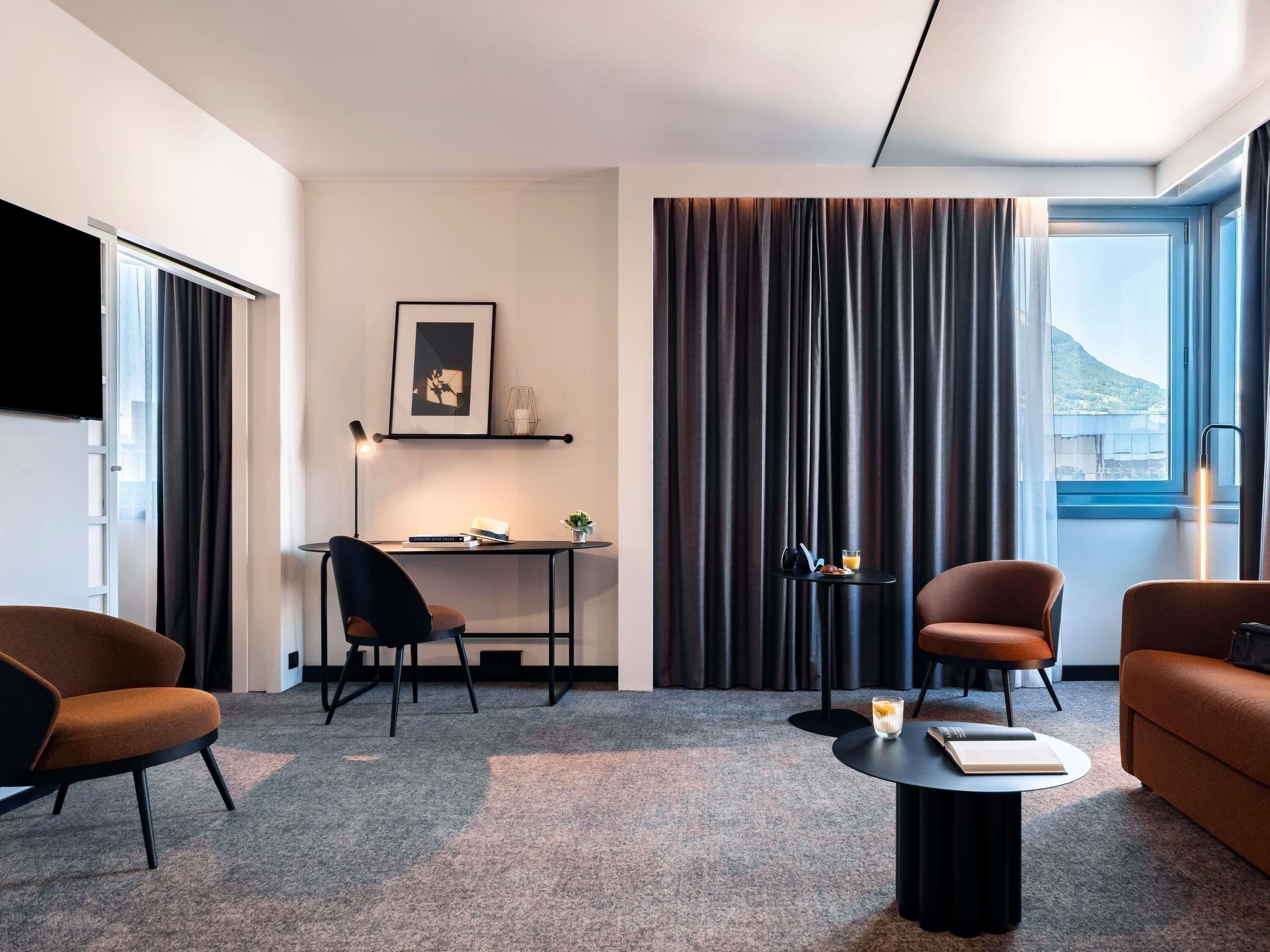 Une chambre d’hôtel moderne équipée d’un bureau, de deux chaises, d’un canapé, d’un téléviseur à écran plat, d’une fenêtre avec des rideaux et d’une étagère murale agrémentée d’un tableau et d’une petite plante.