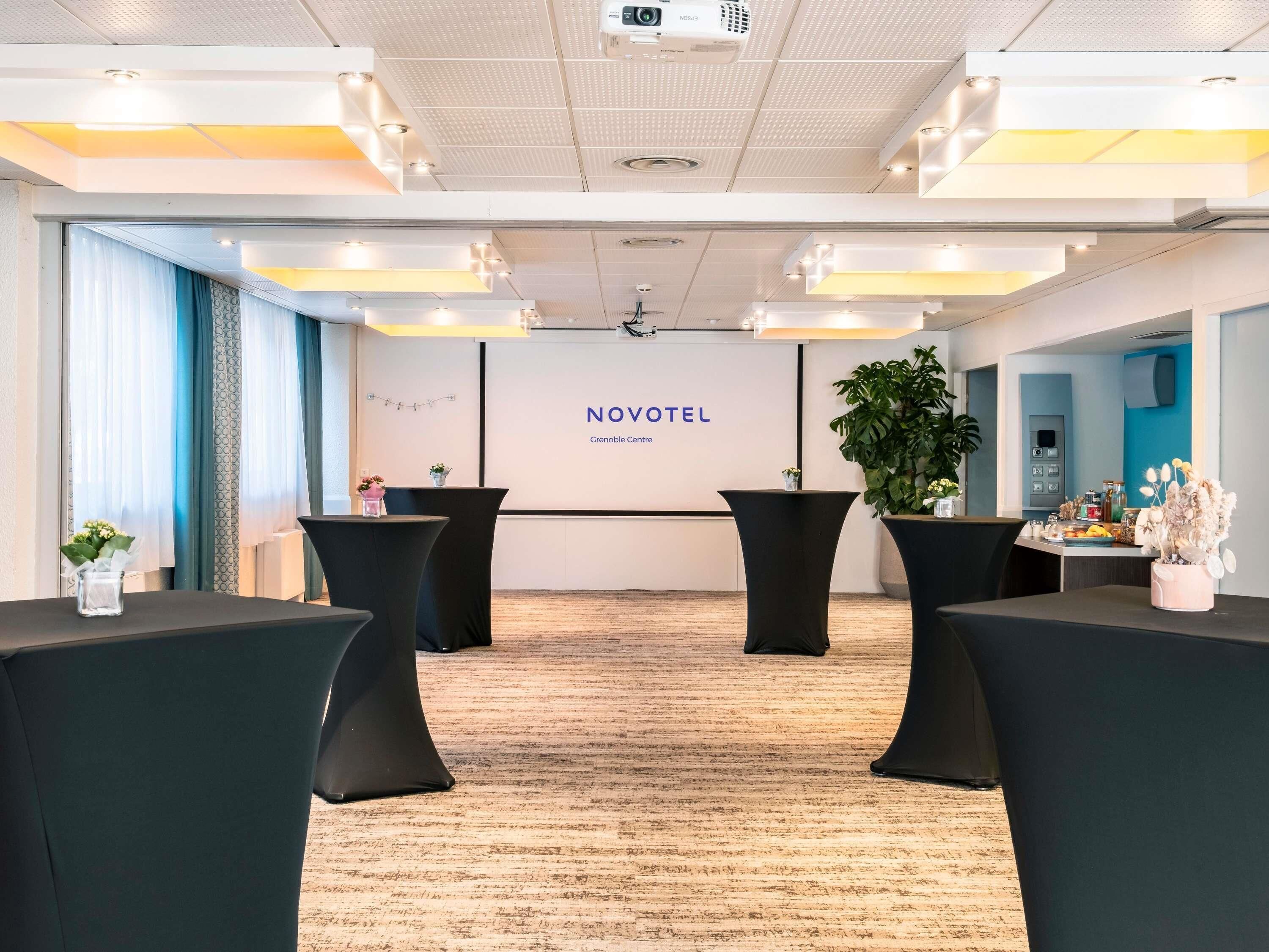 A conference room with a podium, a screen displaying 'NOVOTEL,' and a plant.