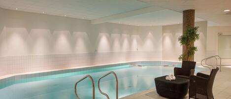 Una piscina interior con paredes de azulejos, agua azul cristalina y una zona de estar con dos sillones de mimbre y un pequeño reposapiés.