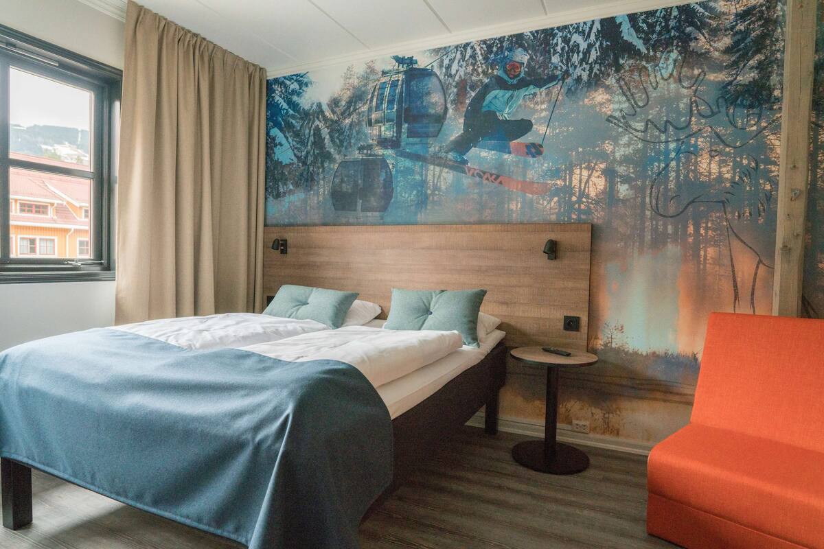 Habitación de hotel con cama, una mesita, una silla, una ventana con cortinas y un mural en la pared.