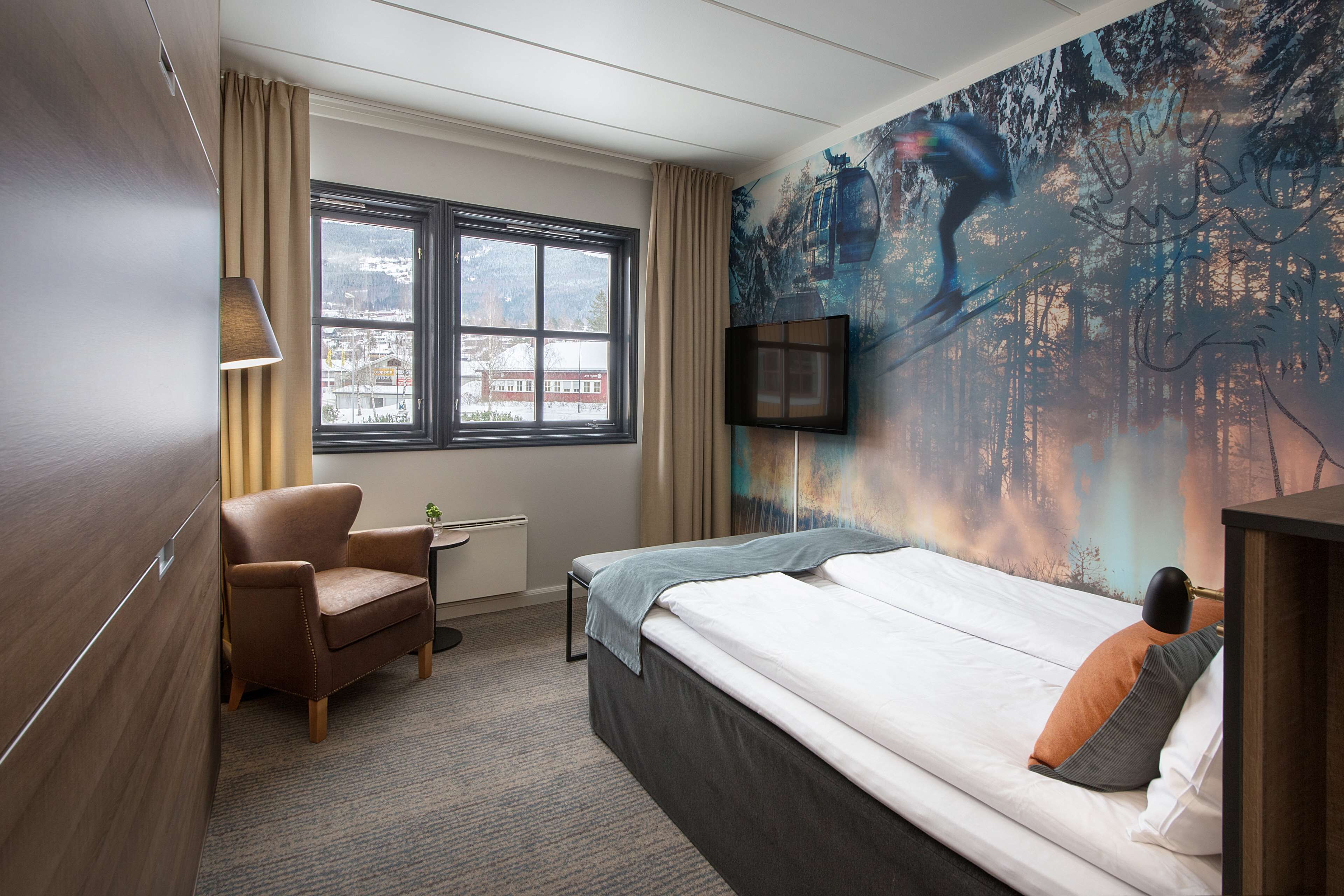 A hotel room with a bed, a chair, a desk, a window with curtains, and a wall mural.