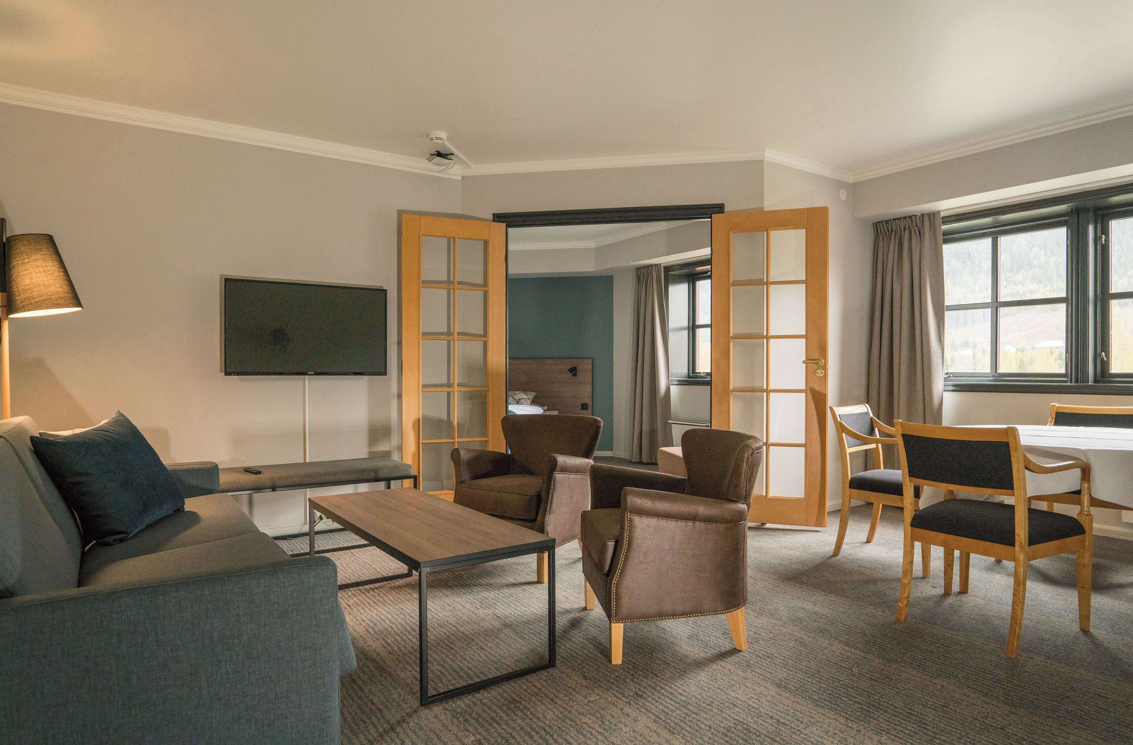 Habitación de hotel con sofá, sillones, una mesa de centro, televisor y un ventanal con cortinas.