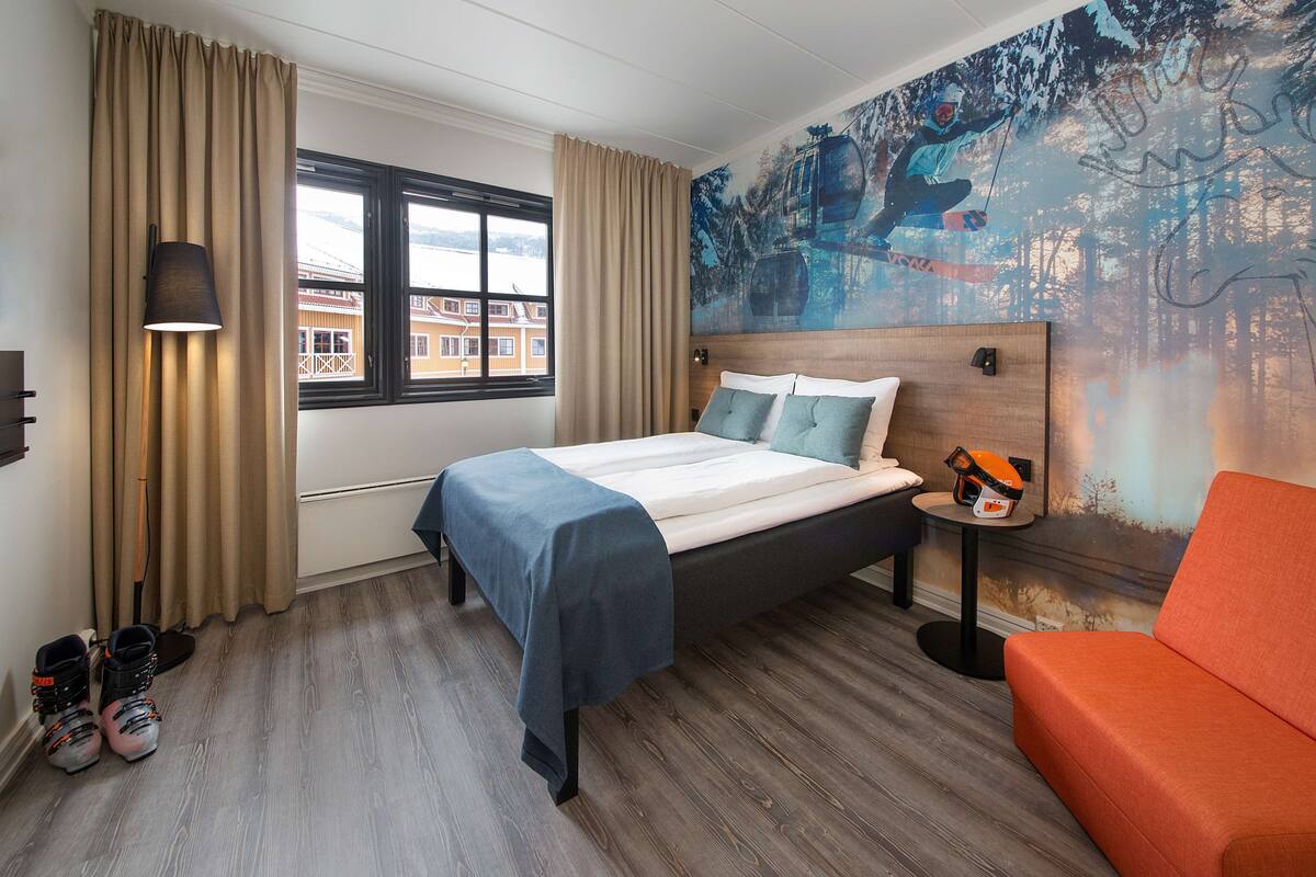 Una habitación de hotel con cama, sofá, ventana con cortinas, lámpara y un cuadro con temática de esquí en la pared.