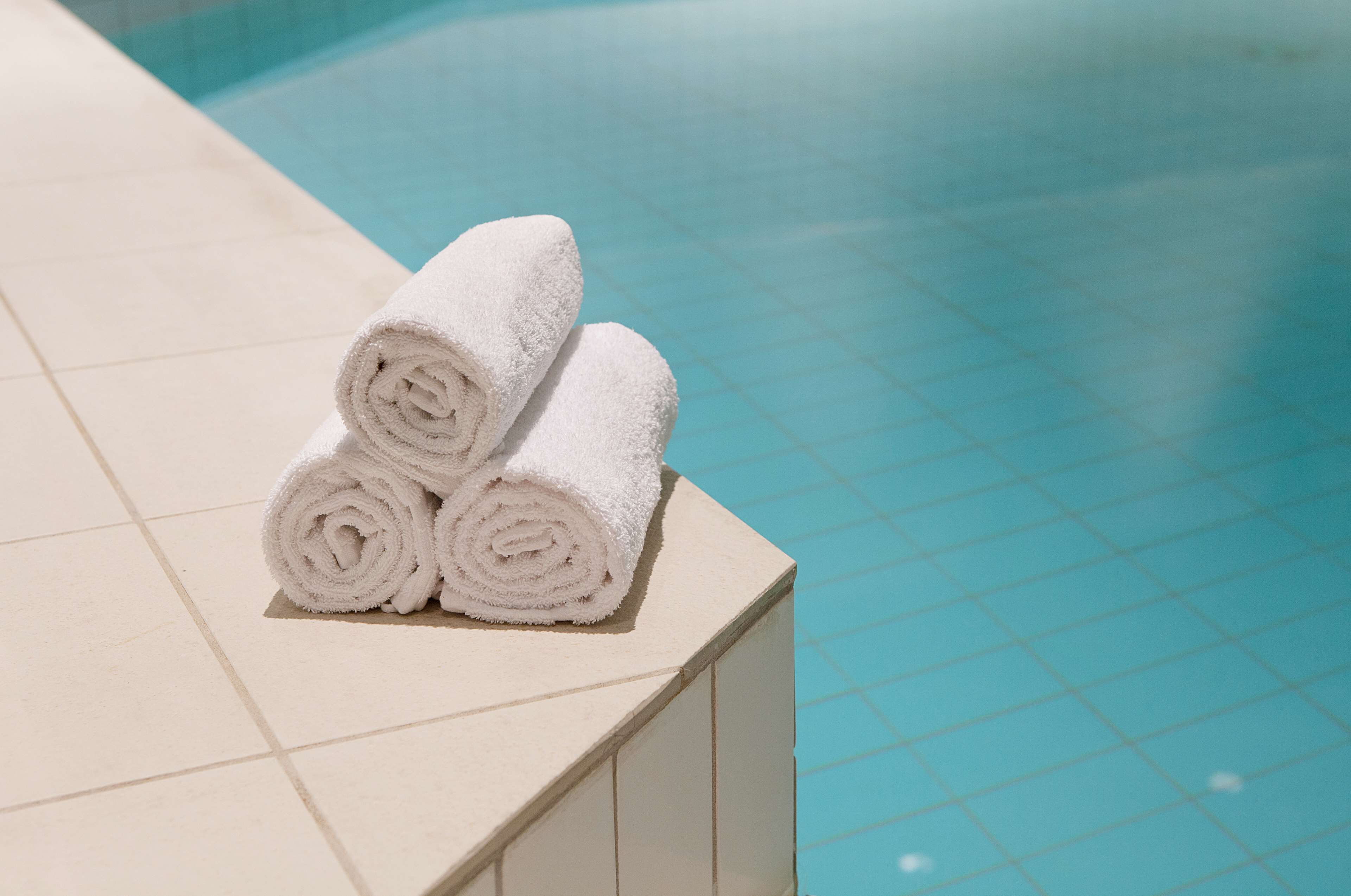 Three rolled white towels stacked on a white tiled ledge by a pool.