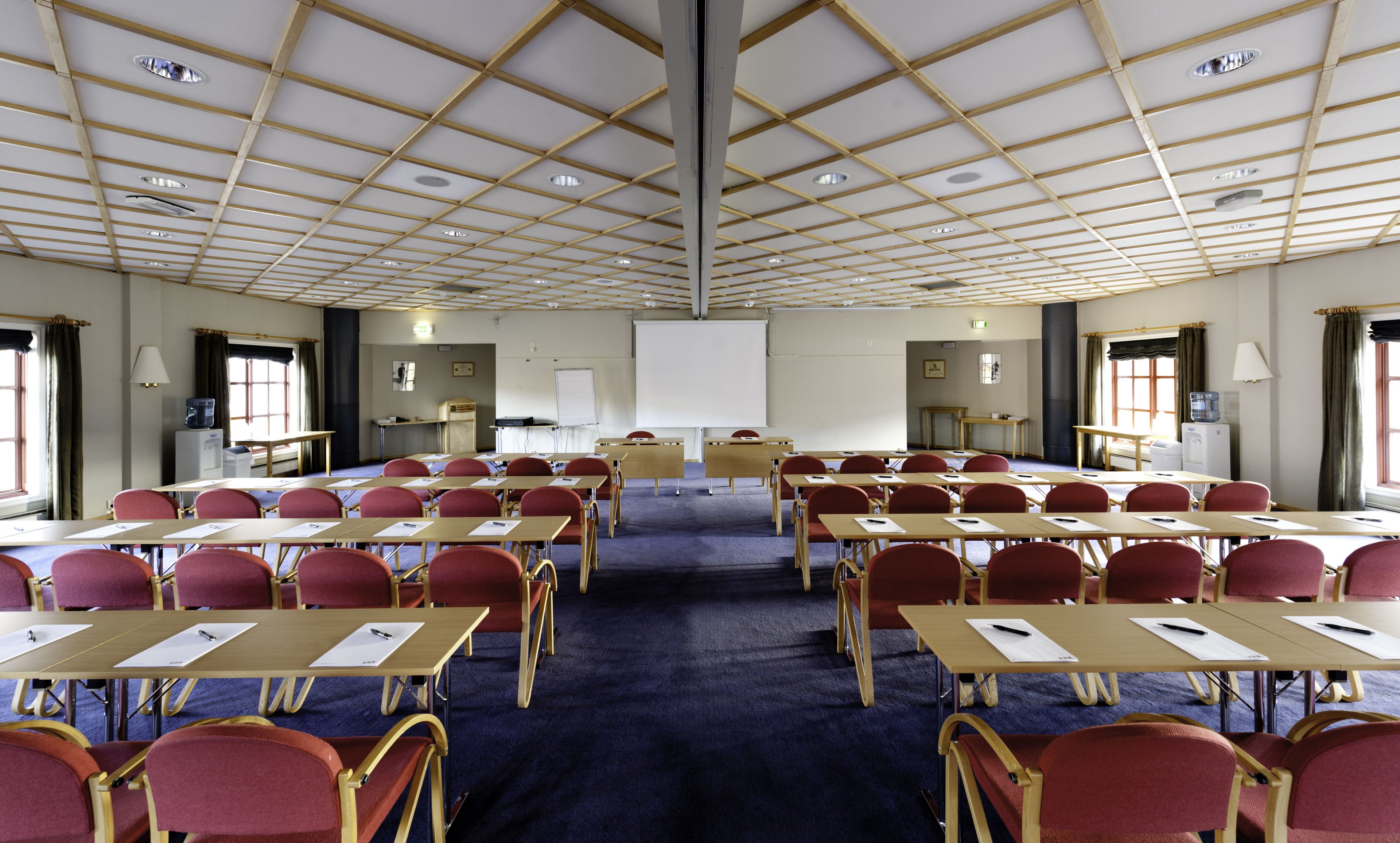 A conference room with a grid-patterned ceiling, multiple tables with chairs, a whiteboard, and a projector screen.