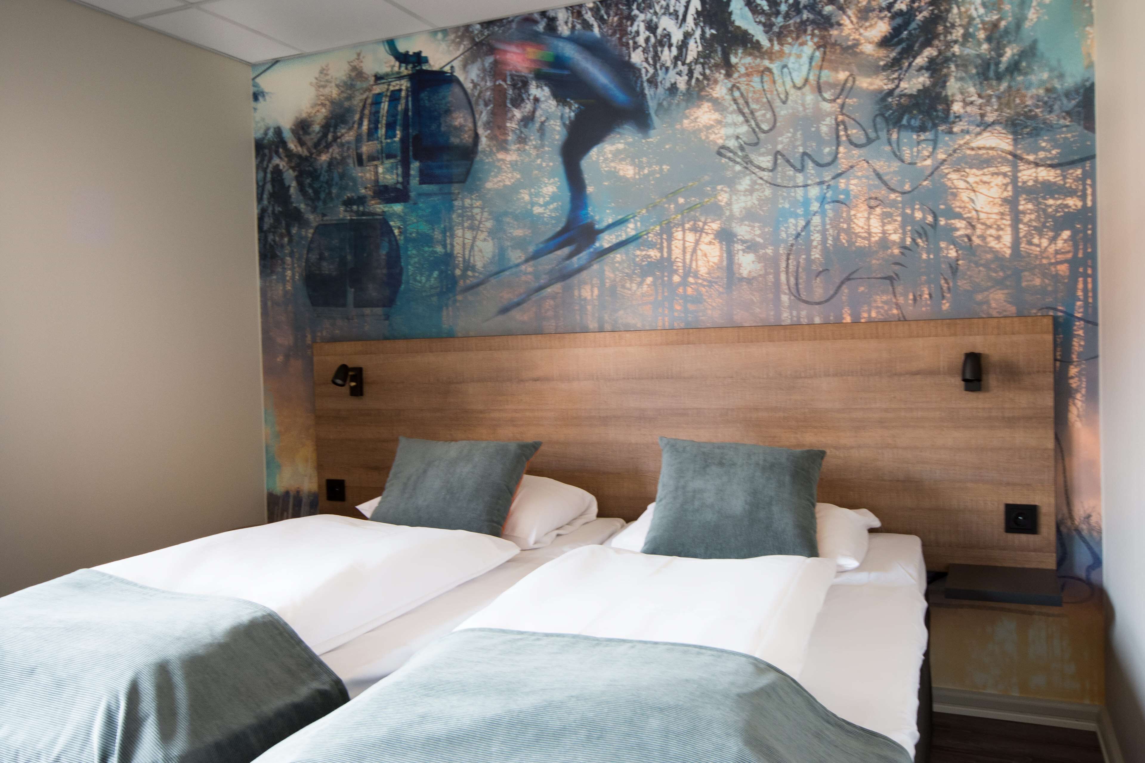 Habitación de hotel con un gran mural de un esquiador en la montaña, cabecero de madera, dos camas con ropa de cama gris y blanca, y una mesita de noche con una lámpara.