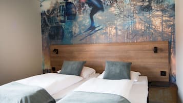 A hotel room with a large mural of a skier on a mountain, a wooden headboard, two beds with grey and white bedding, and a small bedside table with a lamp.
