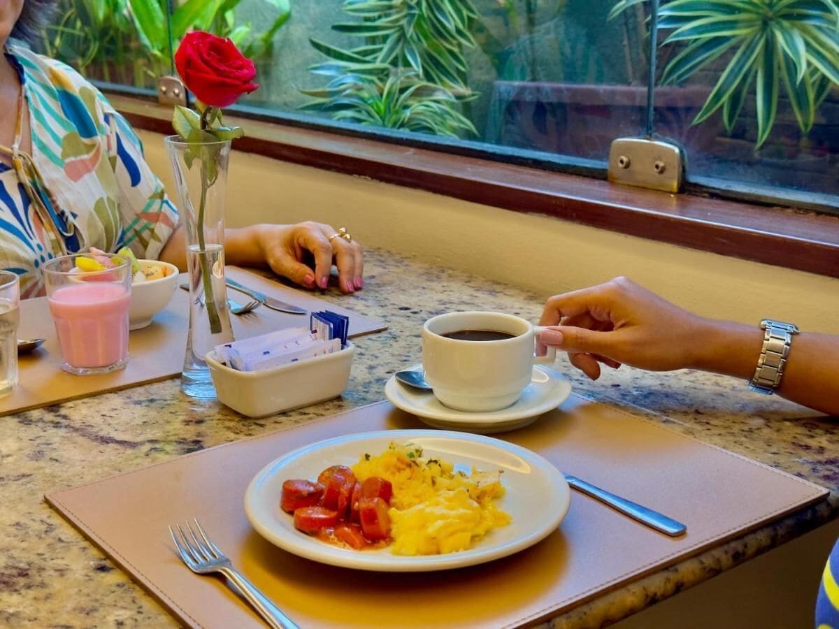 A dining table with a plate of food, a cup of coffee, and a rose in a vase.