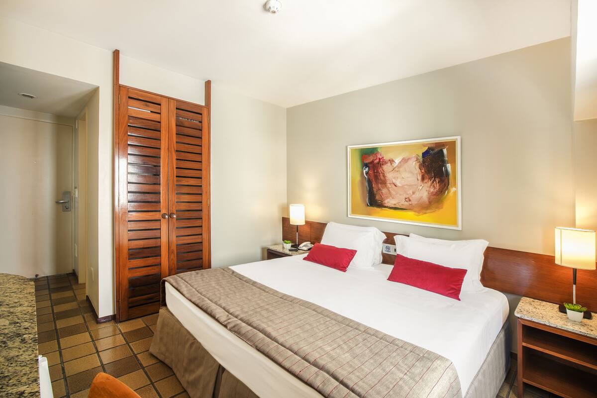 A hotel room with a bed, a wooden wardrobe, a nightstand with a lamp, and a painting on the wall.
