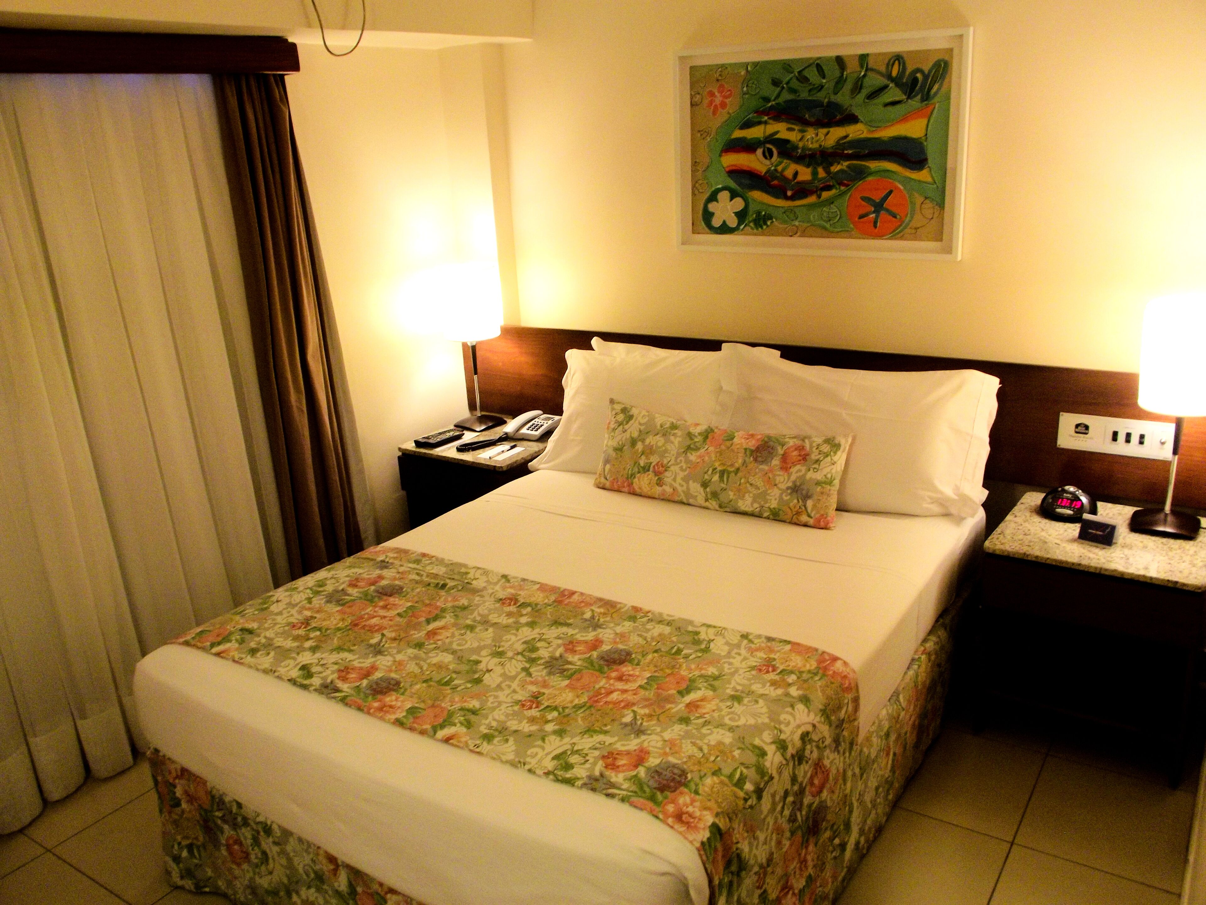 A hotel room with a bed, a nightstand, a lamp, a clock, and a painting on the wall.