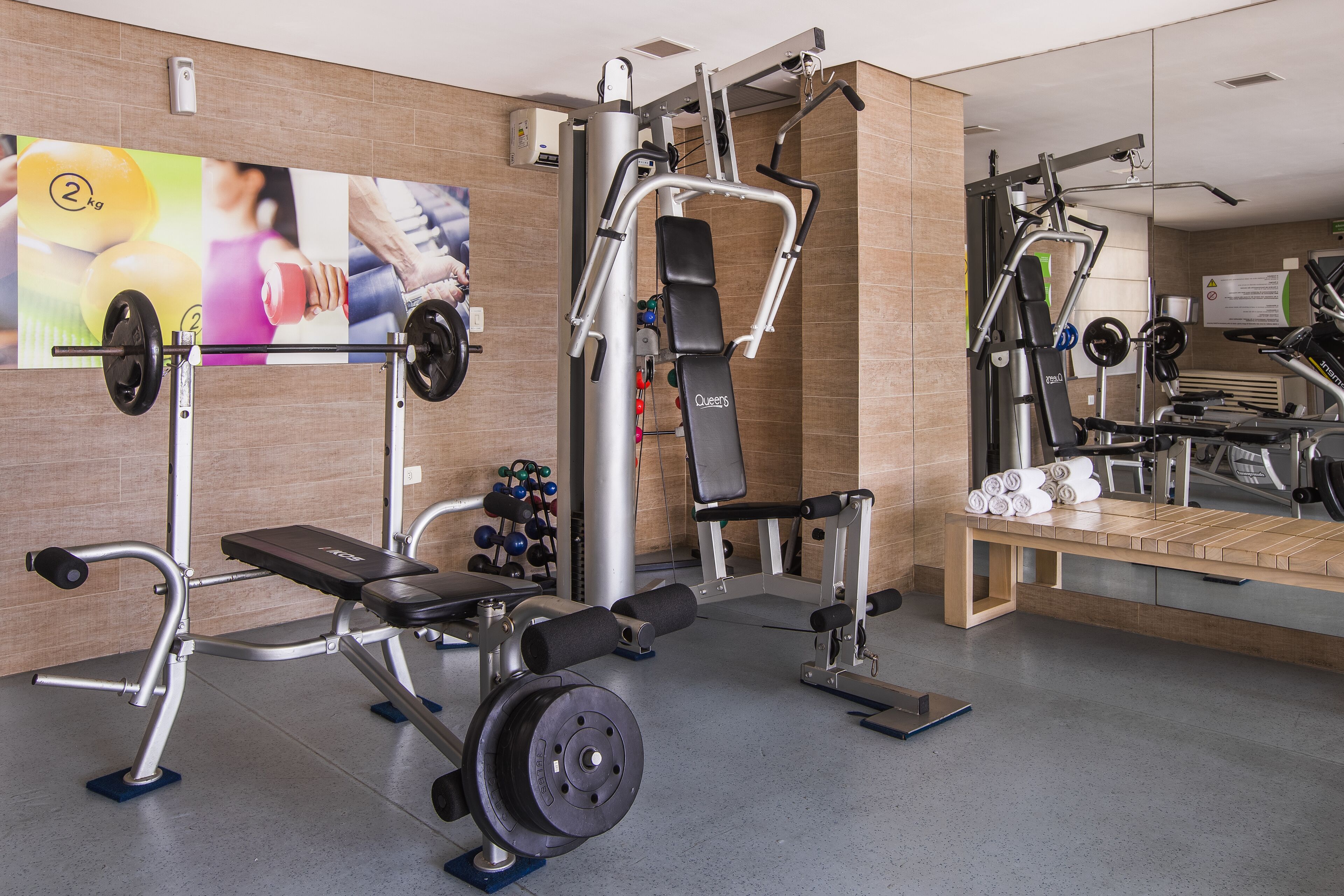 Une salle de sport équipée de divers appareils d’exercice, d’un banc de développé couché et d’un téléviseur fixé au mur qui diffuse du contenu sur le thème du fitness.