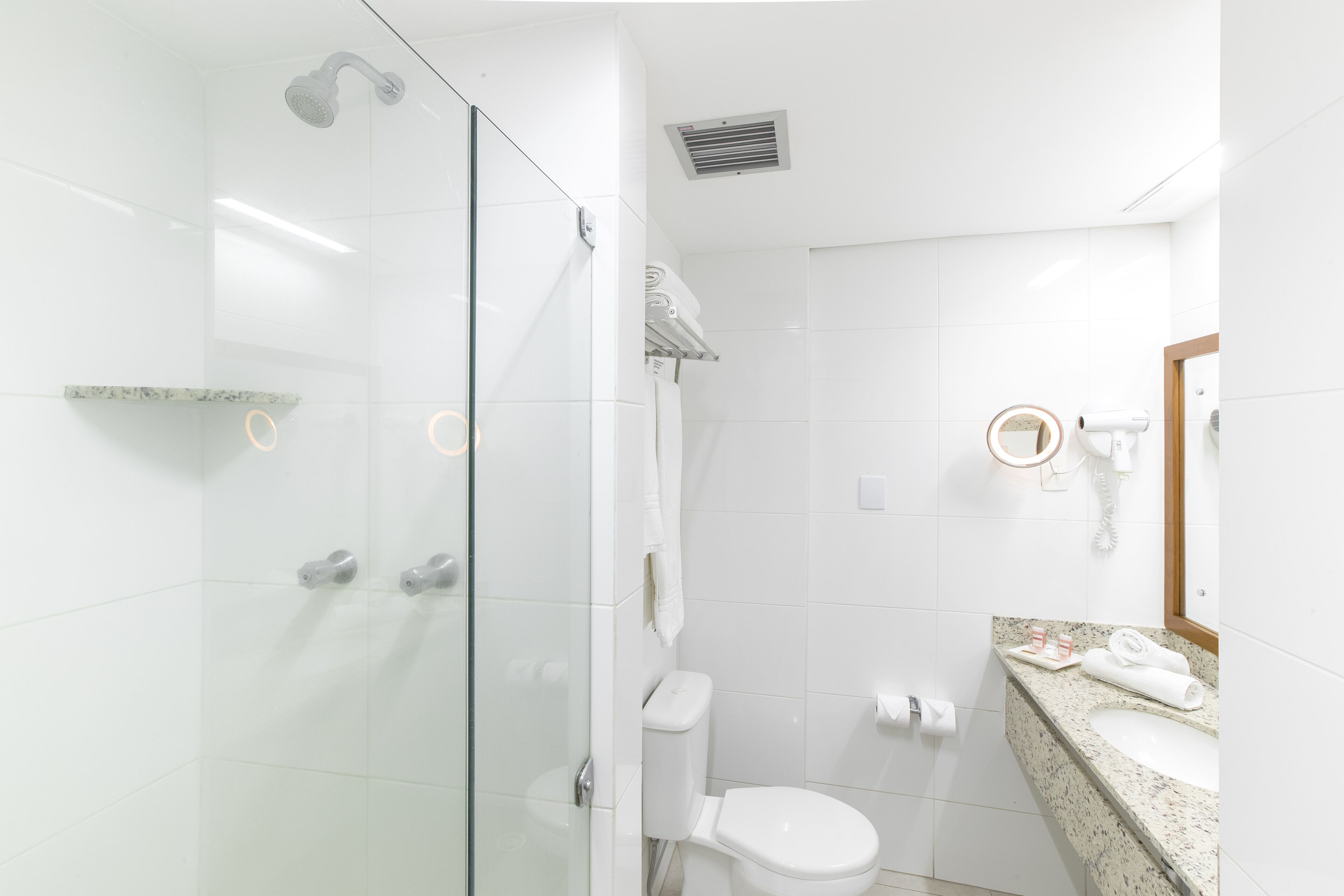 Uma casa de banho moderna com cabine de chuveiro em vidro, sanita branca e bancada de granito com lavatório e porta-toalhas.