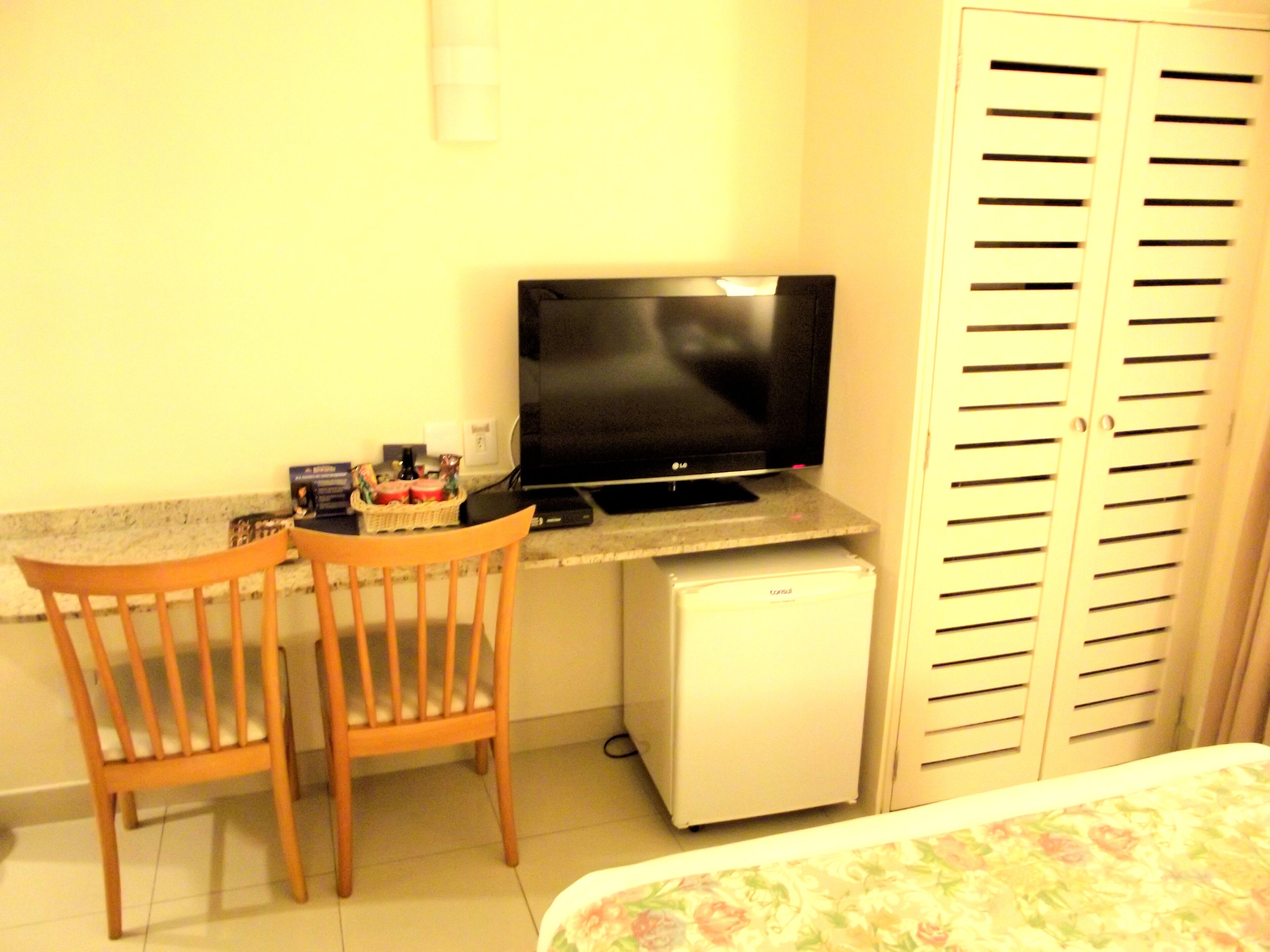 A small room with a desk, a television, a refrigerator, and a wardrobe.