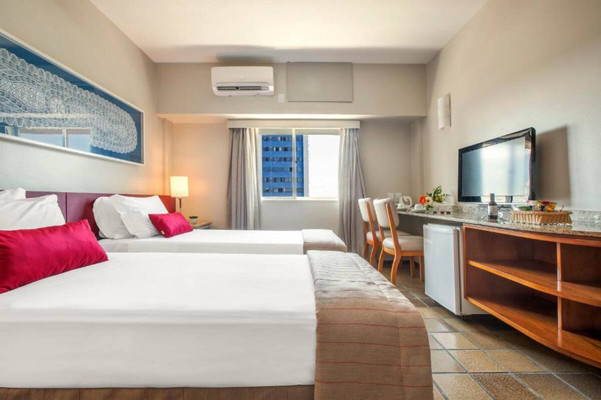 A hotel room with two beds, a desk, a television, and a window with a view of buildings.